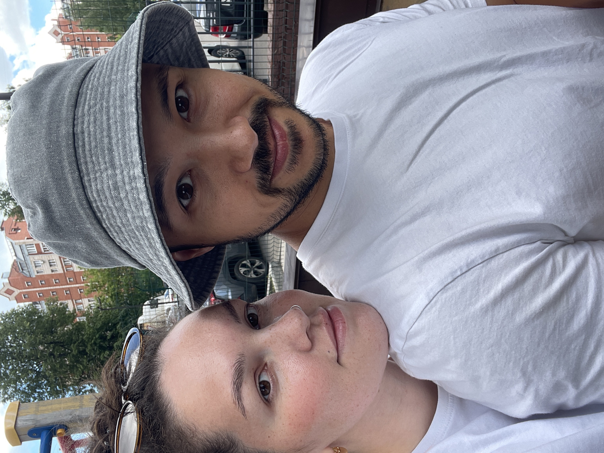 A selfie image captures two individuals outdoors. The male, positioned at the top of the frame, wears a gray bucket hat and a white t-shirt, exhibiting a beard and mustache. The female, located below him, has sunglasses perched on her head, a nose ring, and also dons a white t-shirt. The background reveals an urban setting, complete with buildings, trees, cars and a fence. The scene appears to be a city park or outdoor recreational area. It is impossible to ascertain the exact location in Izhevsk, Russia without additional information.