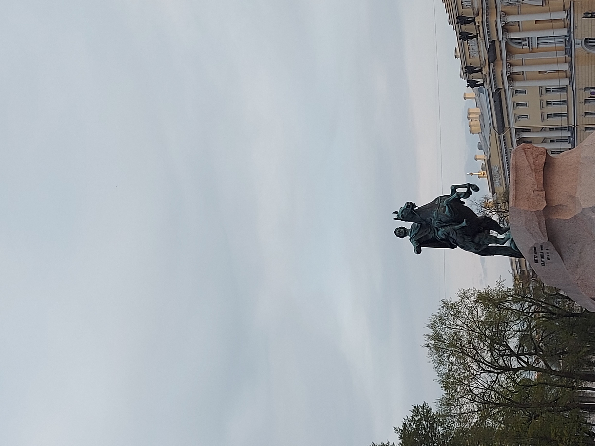 The image shows the Bronze Horseman statue of Peter the Great in Saint Petersburg, Russia. The statue features a rider on horseback, set atop a large natural stone pedestal known as the Thunder Stone. Behind the statue is a pale building with many windows and columns. Below the statue, are bare trees. The sky is a uniform grey color.