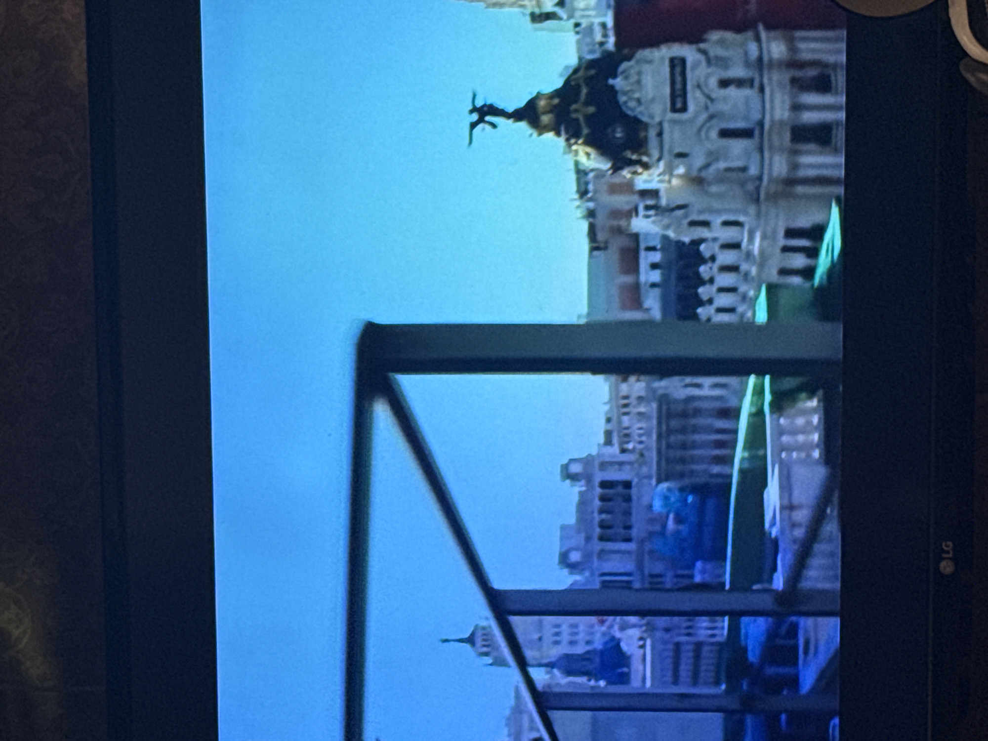 The image shows a view of the city of Madrid, Spain, seen through what appears to be a window frame. The building with the statue on top appears to be the Metropolis Building in Madrid. Other buildings are visible in the background, under a clear blue sky. The photo is a tilted perspective. The image is on a screen with the LG logo on it.