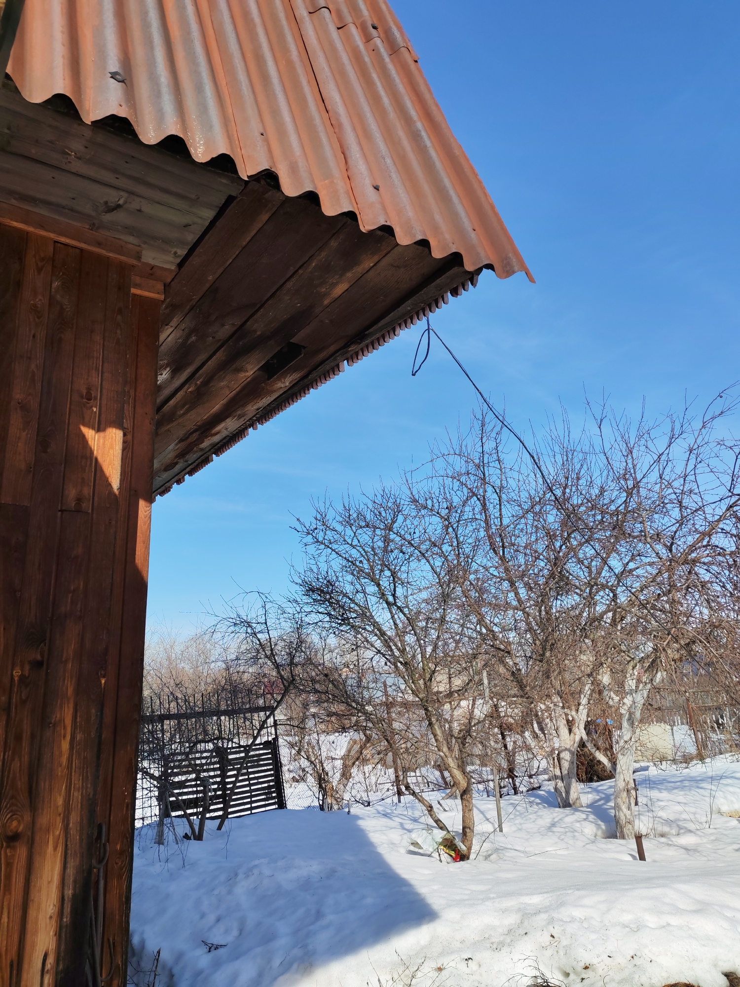 The image presents an outdoor scene in what appears to be a rural or semi-rural setting. Patches of snow cover the ground, suggesting a winter or early spring season. In the foreground, a portion of a wooden structure with a rusty, corrugated metal roof is visible. The wood has a dark, aged appearance, and the roof shows signs of weathering.
Beyond the structure, there are several leafless trees and bushes, indicating dormancy typical of colder climates. A metal fence is partially visible in the mid-ground, suggesting property boundaries. The background features a clear, blue sky. There is no direct evidence to confirm the location as Chaikovsky, Russia, or to identify any people, specific actions, or interactions. The scene predominantly depicts a still, wintry landscape with a focus on the architecture and natural environment.