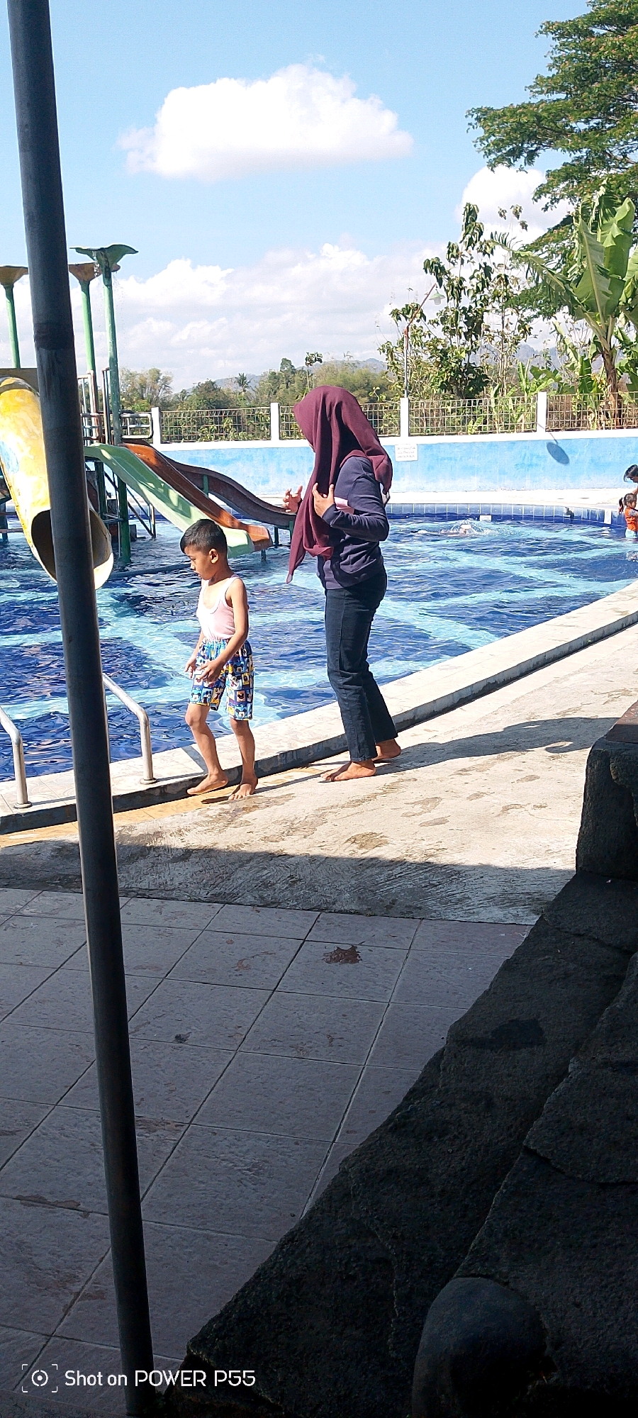 A medium shot captures a bright, sunny day at an outdoor swimming pool. Two people are visible near the edge of the pool. On the left, a young boy with short dark hair, wearing a light pink tank top and patterned swimming trunks, stands at the pool's edge, looking down. To his right, a person wearing a deep purple hijab and dark blue long-sleeved shirt and pants, stands with their back to the camera, facing the pool. They appear to be interacting with the boy, possibly guiding or talking to him. The swimming pool is filled with clear blue water, with white lane markers visible. To the left of the frame, a yellow and green water slide is partially visible. The background shows a tiled walkway, a white fence, lush green trees, and a hazy, mountainous landscape under a bright blue sky with fluffy white clouds. The overall scene suggests a leisurely day at a public or private swimming facility.