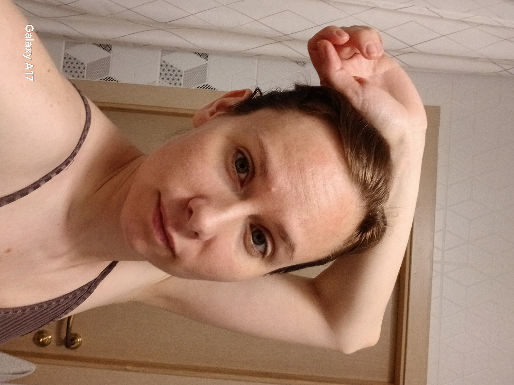 A woman with light skin and short brown hair poses for a selfie. Her right arm is raised above her head. She wears a brown tank top. The background includes a white patterned wall and a light brown door with gold hardware. The text "Galaxy A17" is visible in the upper left corner of the image. The location is not explicitly identifiable as Yekaterinburg, Russia.