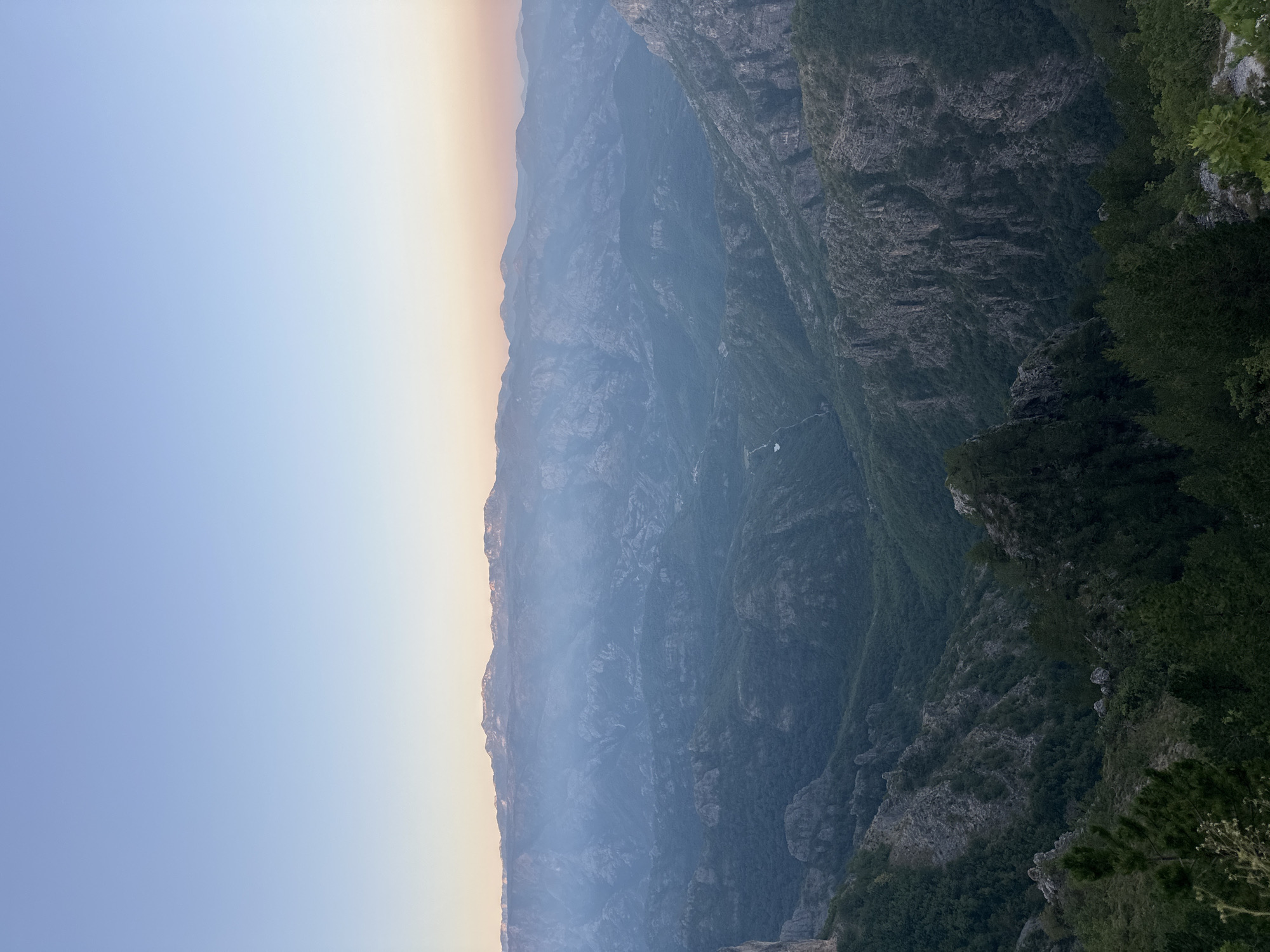 This image captures a panoramic view of a mountainous landscape at dusk. The sky transitions from a pale blue at the top to a soft orange and pink hue near the horizon, suggesting the setting sun. Below the sky, layers of mountains recede into the distance, with the closest peaks appearing dark green and densely forested, while those further away are shrouded in a light blue haze, indicating atmospheric perspective. The terrain is rugged, characterized by steep slopes, rocky outcrops, and deep valleys. A narrow, winding path or a dry riverbed can be observed in one of the valleys. The foreground features the tops of dark green pine trees, adding depth to the scene. The overall impression is one of natural grandeur and tranquility.
The location of Aranđelovo, Bosnia and Herzegovina, is not explicitly identifiable from the visual elements present in the image. The scene depicts a generic mountainous region.