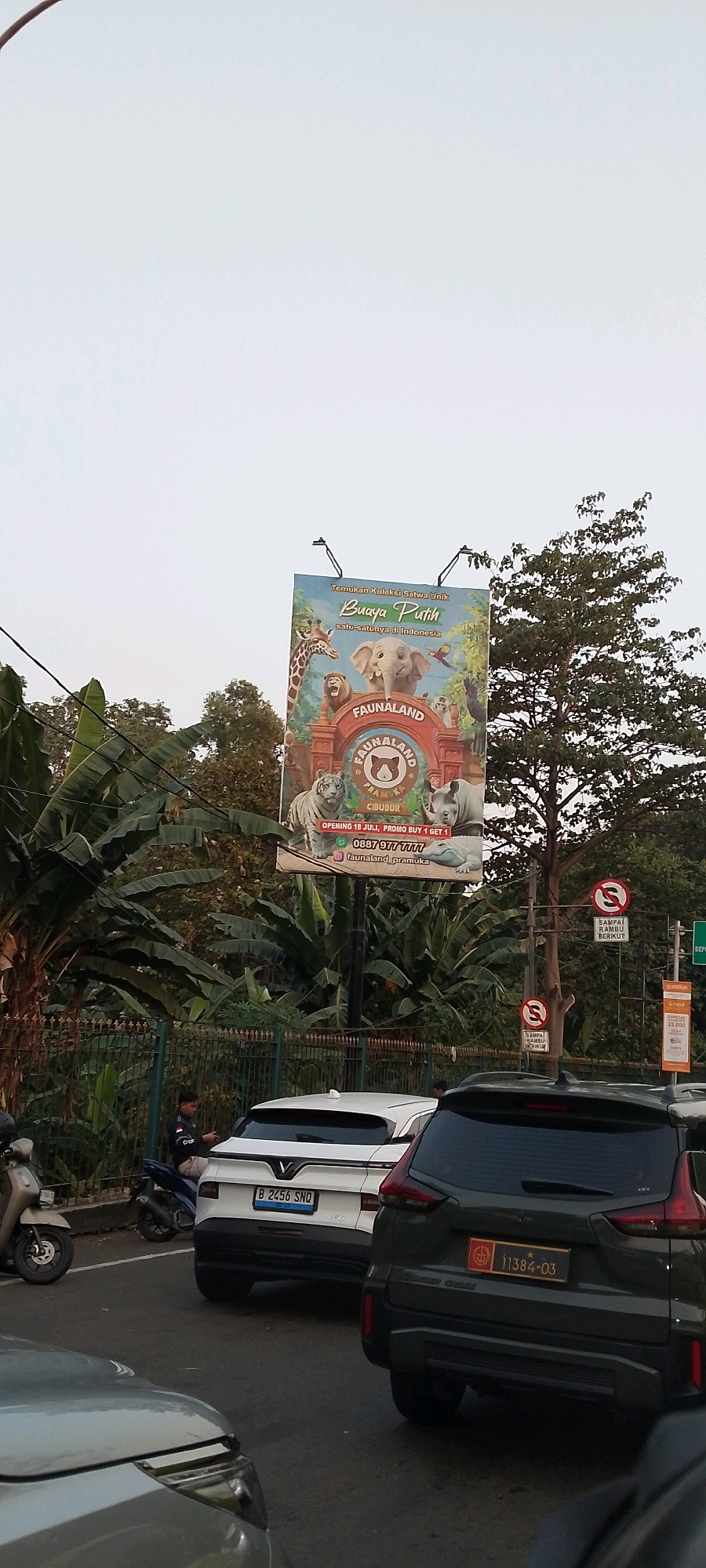 The image captures an outdoor scene, likely in East Jakarta, Indonesia, featuring a large billboard for "Faunaland" zoo. The billboard displays various animals like a giraffe, elephant, tiger, rhino, and a crocodile, along with the text "Buaya Putih" (White Crocodile) and "satu-satunya di Indonesia" (the only one in Indonesia). It also mentions "OPENING 18 JULI, PROMO BUY 1 GET 1" and a phone number "0887 977 7777" and social media handle "@faunaland_pramuka".
In the foreground, several vehicles are visible, including a white SUV with license plate "B 2456 SNQ" and a dark green SUV with license plate "11384-03". A person is seated on a motorcycle near the white SUV. The environment includes lush greenery with large banana leaves and trees, a metal fence, and traffic signs indicating restrictions. The sky is overcast. The overall scene suggests a street view near the zoo entrance or an advertising location.