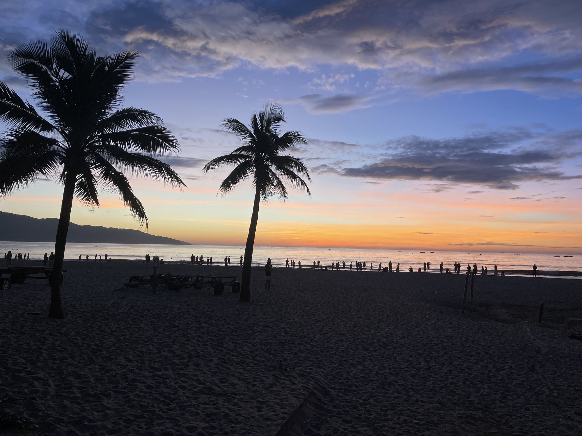 The image depicts a beach scene at dusk, likely on Mỹ Khê Beach in Đà Nẵng, Vietnam. Silhouetted palm trees dominate the foreground, with their fronds stark against the twilight sky. The sky displays a gradient of colors, from deep blue and purple at the top to vibrant oranges and pinks near the horizon, indicating the sun has recently set. A calm ocean stretches out, meeting a distant, dark shoreline or mountain range. Along the water's edge, numerous people are visible as dark silhouettes, scattered across the sand and wading in the shallow water, suggesting a leisurely evening activity. Several small boats are also present on the water. The beach itself is sandy, with footprints and subtle undulations marking the surface. In the middle ground, two dark, wheeled structures, possibly carts or beach equipment, are placed on the sand. The overall atmosphere is serene and picturesque, characteristic of a tropical beach sunset.