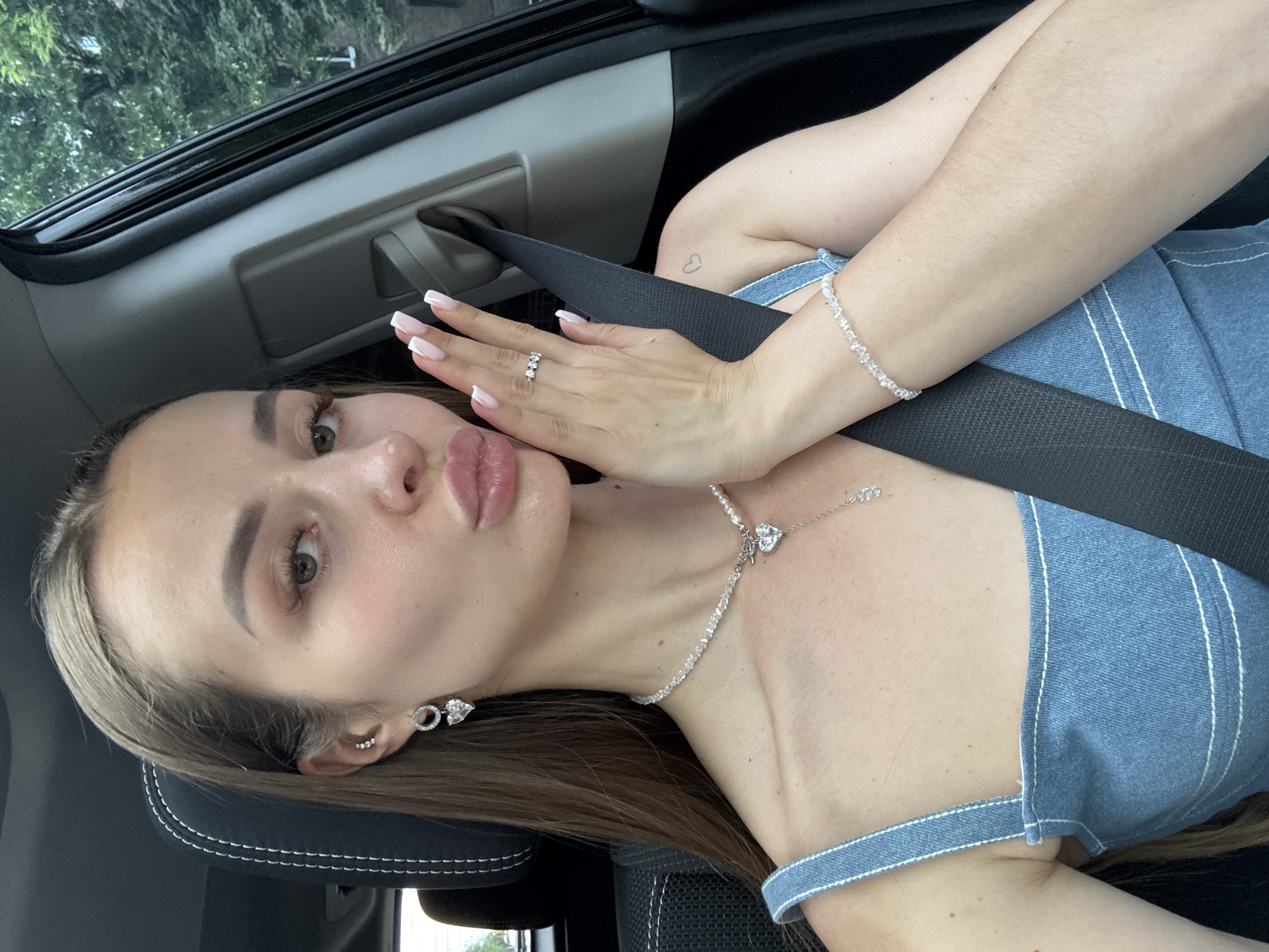 A young woman with long, brown hair is taking a selfie in the passenger seat of a car. She is wearing a denim top and a necklace with a heart pendant. She has on earrings, a ring, and a bracelet. Her left hand is resting on her cheek, and she is pursing her lips. The interior of the car is visible, including a black seatbelt. The background outside the window is blurred, showing green trees, suggesting an outdoor setting, possibly in Kyiv, Ukraine. The overall mood is casual and playful.