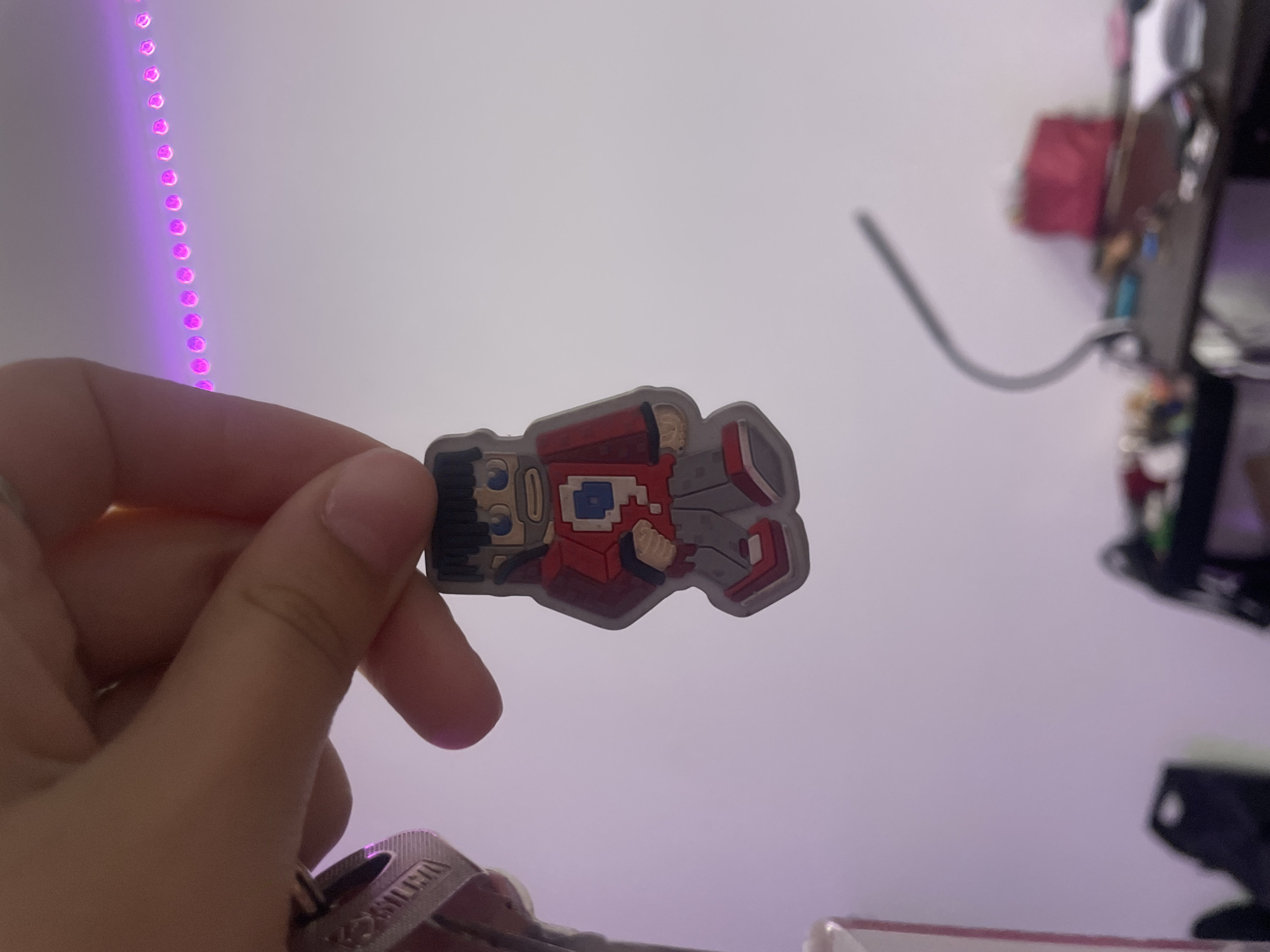 A person's hand holds a small, blocky figurine with a red jacket and gray pants. The figurine resembles a character from a video game. In the background, a string of pink LED lights runs horizontally along the wall. The scene is indoors, and the lighting suggests it is nighttime or a dimly lit room. The location of Лесное Снт, Russia is not depicted or inferable from the image.