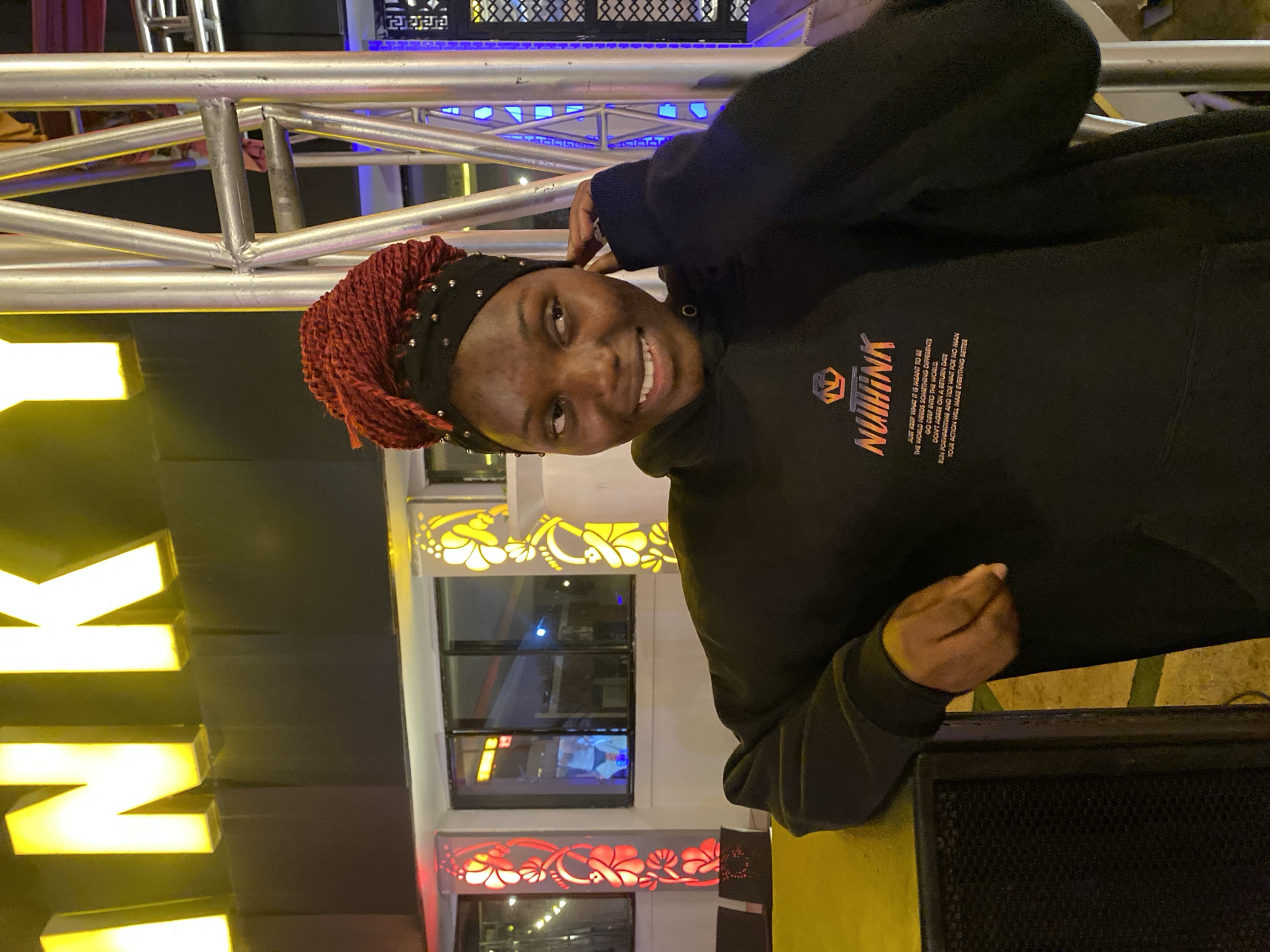 A young Black woman with braided red hair and a black headscarf is leaning against a dark speaker in front of a building with bright yellow signs and decorative pillars. She is wearing a black hooded sweatshirt with a graphic and text on it. Behind her, there is a metal structure and some lights. The overall setting appears to be an outdoor entertainment venue.