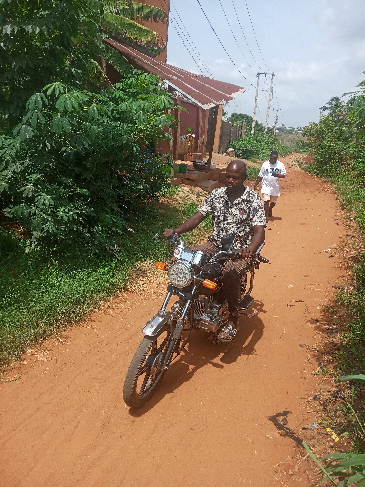 The image captures a daytime scene on a reddish-brown dirt road in Ukwu-Abwa, Nigeria. In the foreground, a bald man is riding a motorcycle, moving towards the right. He wears a patterned short-sleeve shirt, dark trousers, and Adidas slides, with a shoulder bag across his chest. The motorcycle features chrome accents, a black seat, and a round headlight with multiple LED lights. The brand name "KYMSTONE" is visible on the front of the motorcycle below the headlight.
The unpaved road is bordered by dense green vegetation, including noticeable cassava plants on the left. In the mid-ground, another man walks away from the viewer along the same dirt path, dressed in a white t-shirt and light-colored shorts. Further back on the left, near a simple building with a rusty corrugated iron roof, a child in a yellow top and dark bottoms is standing next to a basin or bucket.
The background reveals more greenery, additional buildings, and utility poles with overhead power lines stretching across a bright, partly cloudy sky. Strong sunlight casts clear shadows, suggesting it is midday or early afternoon. The scene depicts typical daily activity in a rural or semi-urban setting.