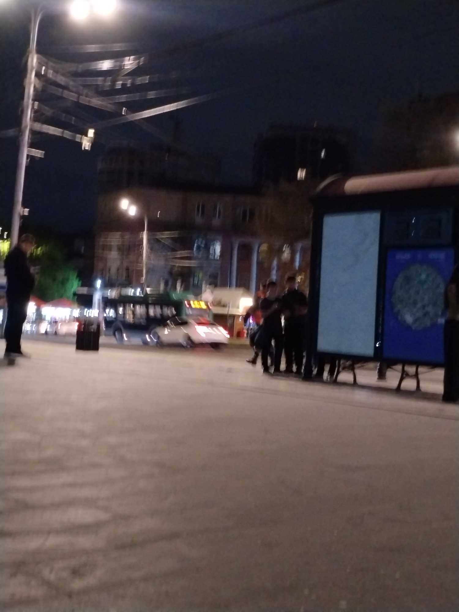 The image depicts an urban street scene at night in Yerevan, Armenia, characterized by artificial lighting and a general blurriness. The foreground shows a light-colored paved surface, likely a public square or sidewalk.
On the left, a solitary individual in dark clothing stands, partially illuminated. In the mid-ground, a dark bus and at least two white cars are visible, suggesting traffic or a public transport area. A dark crate is situated on the ground in this section.
To the right, a group of four to five people, predominantly in dark attire, are gathered near two large, illuminated vertical signs. One sign is mostly light blue/white, and the other is dark blue with a circular graphic, possibly a map or information display, though no discernible text is legible due to the blur. Another person is partially visible on the far right.
Blurry buildings with some illuminated windows form the backdrop. Overhead, bright streetlights are prominent, casting a glow and creating glare. A network of metal structures, possibly for utility lines like trolleybuses or decorative lighting, crisscrosses above. The dark sky confirms the nighttime setting. The overall scene suggests a casual evening gathering or transit activity in a city environment.