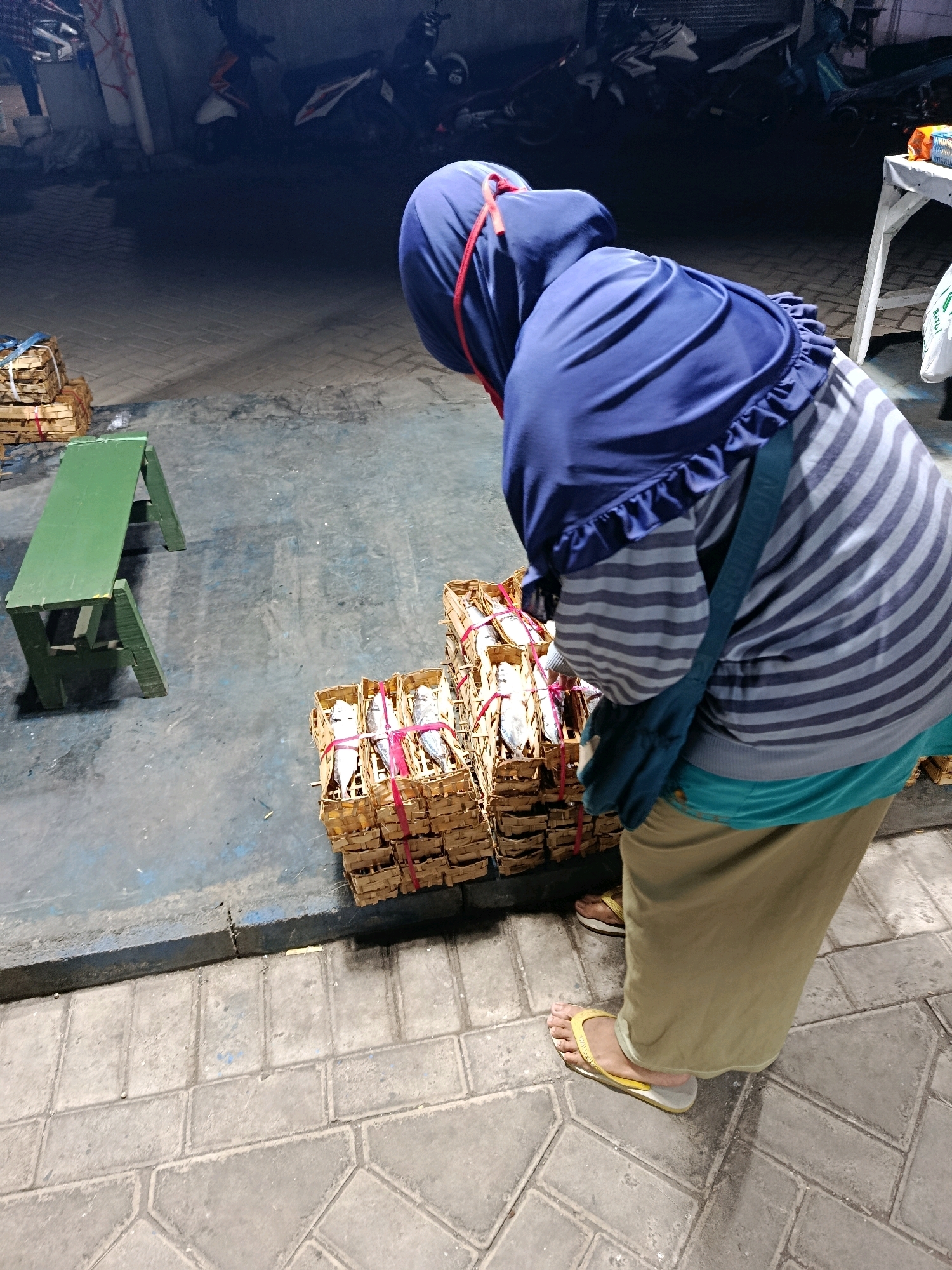 In Malang, Indonesia, at night, a person wearing a blue hijab and a striped shirt bends over small woven baskets filled with fish. The person also wears a tan skirt and yellow flip-flops. In the background, motorcycles are parked. There is a green bench to the left of the image. The ground is paved with stones and asphalt.