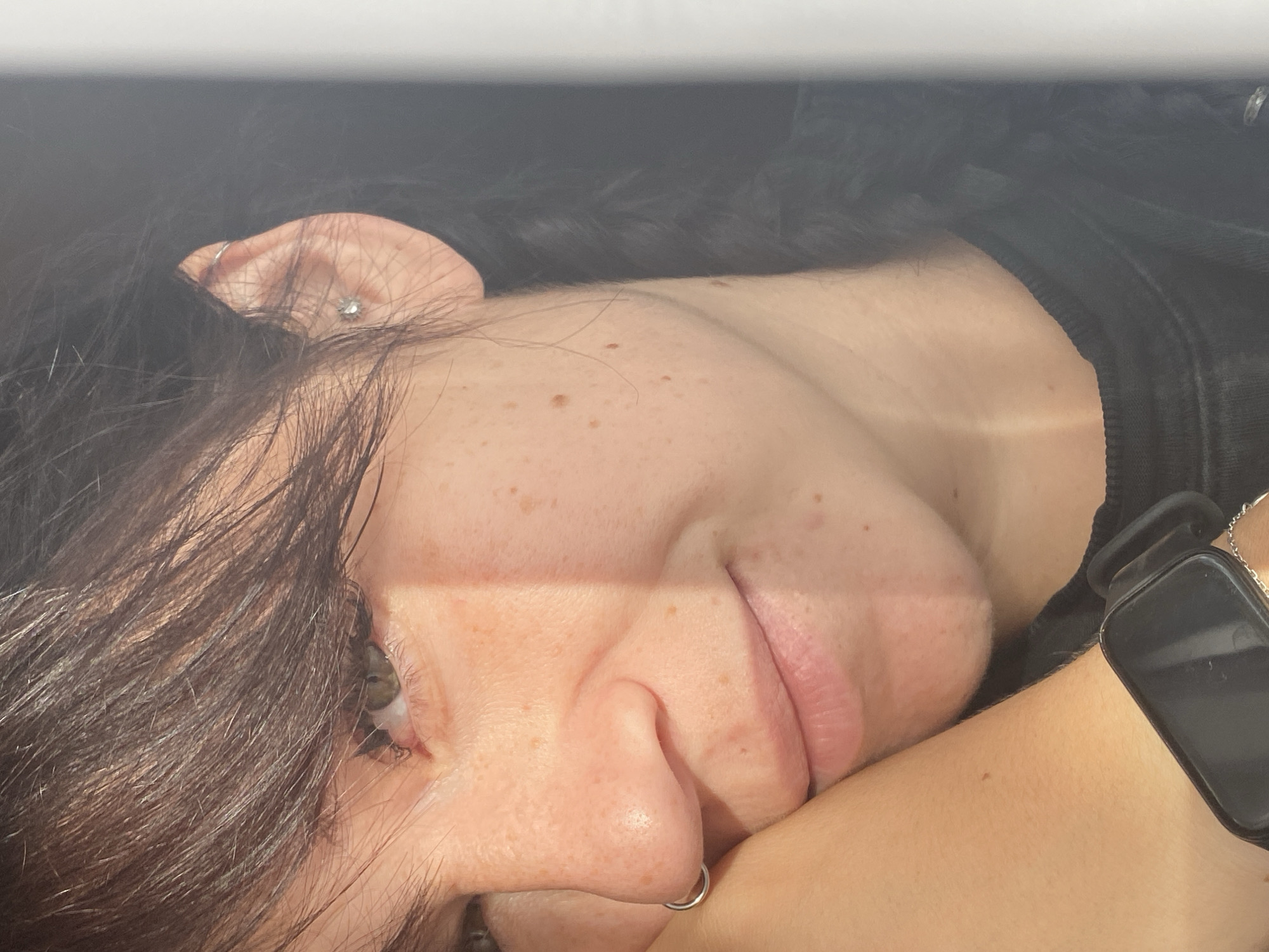 The close-up, vertical, slightly-above-eye-level shot showcases the left side of a person's face. The subject's dark hair is styled with bangs falling across their forehead, and a braid extends down their back. They have green eyes, a nose with a silver hoop piercing, and a gentle smile. Their left arm rests beneath their chin, and a smartwatch is visible on their wrist. The skin has freckles and is illuminated by a strong light source, creating a distinct shadow line across the face. The background is dark and blurred, with a bright vertical element on the right edge of the frame. The location is identified as Krasnoflotskoe, Russia.