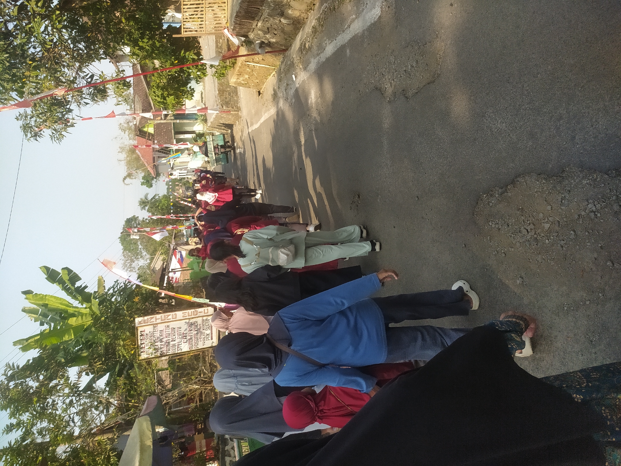 The image depicts a street scene in Lopait, Indonesia, likely during a local event or procession. A group of people, predominantly women and children, are walking along a paved road. Many are wearing headscarves, with some in matching maroon shirts, suggesting a group activity or celebration.
In the foreground, a person in a blue shirt and dark pants is walking away from the camera, followed by a young child in a red headscarf. To the right, a woman in a light green outfit with a backpack is walking in the same direction.
Along the side of the road, there are houses with red-tiled roofs and some vegetation, including banana trees and other greenery. Red and white flags are strung along poles, indicating a festive or patriotic occasion. A sign is visible on the left, written in Indonesian, appearing to list services or goods. On the right side of the road, there is a stone wall and a wooden fence. A motorbike with a large basket on the back is also present, carrying what appears to be goods. The lighting suggests it is daytime, with shadows cast on the road.