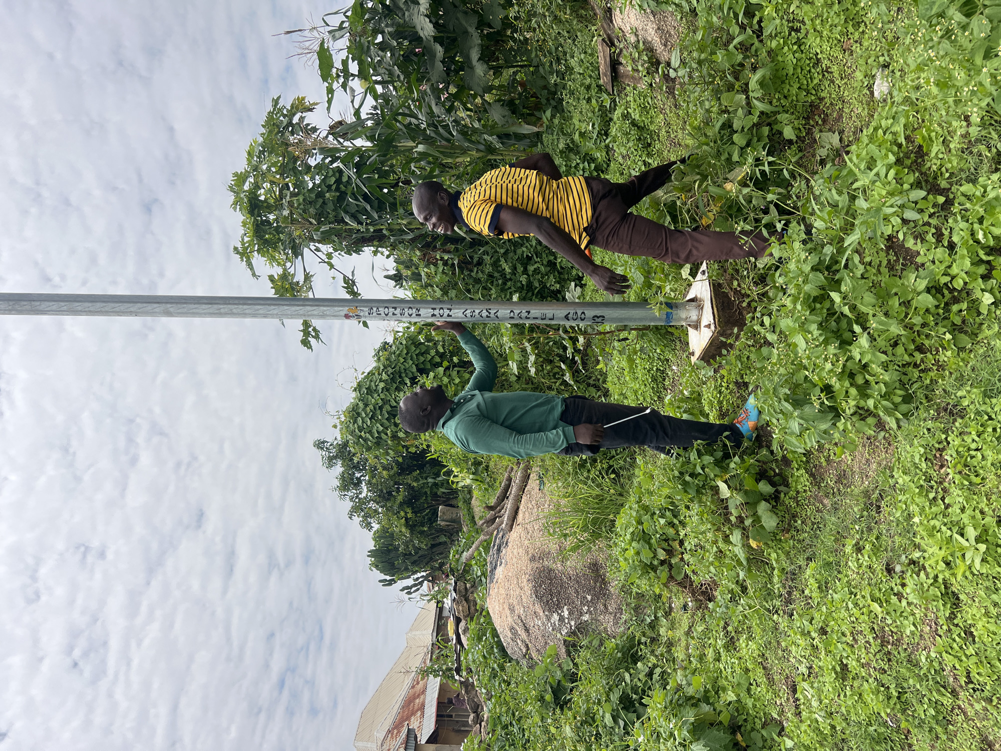 Two men stand near a metal pole in a grassy, overgrown area. The man on the left, wearing a green long-sleeved shirt and dark pants, is touching the pole. The man on the right, wearing a yellow and black striped shirt and brown pants, is smiling and looking upwards. The pole has text printed vertically on it, including "SPONSOR HON ASAMA DANIEL AGO". The background features lush vegetation, including corn stalks, and a building with a corrugated metal roof. The scene is set outdoors with a cloudy sky. This image was taken in Dong, Nigeria.