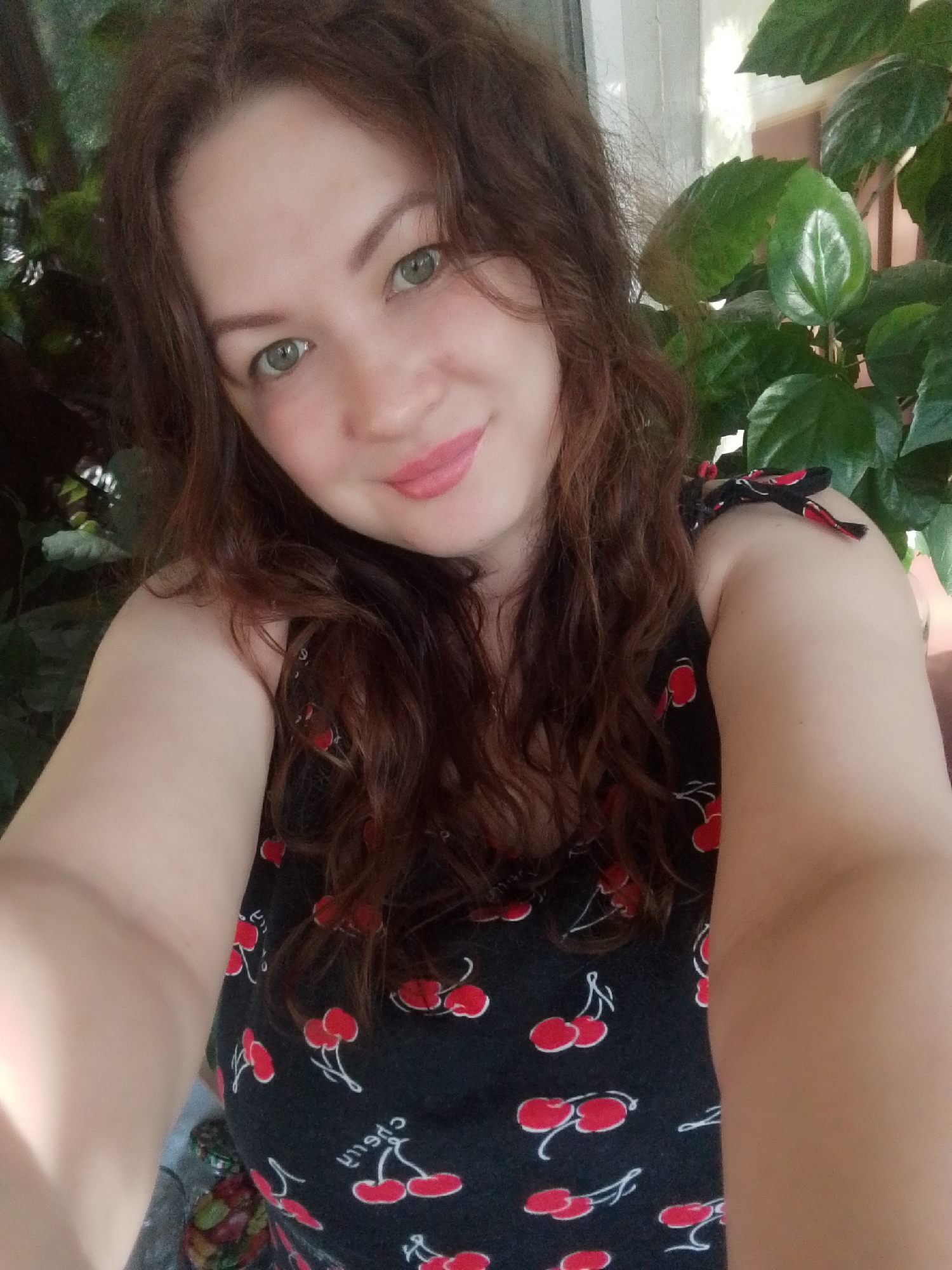 A woman with fair skin, light green eyes, and curly brown hair is taking a selfie. She is wearing a black sleeveless top with a cherry print. She has pink lipstick on. The background includes green leafy plants. The location is Noviy, Russia. The specific environment includes the presence of plants, possibly indoors or on a balcony.