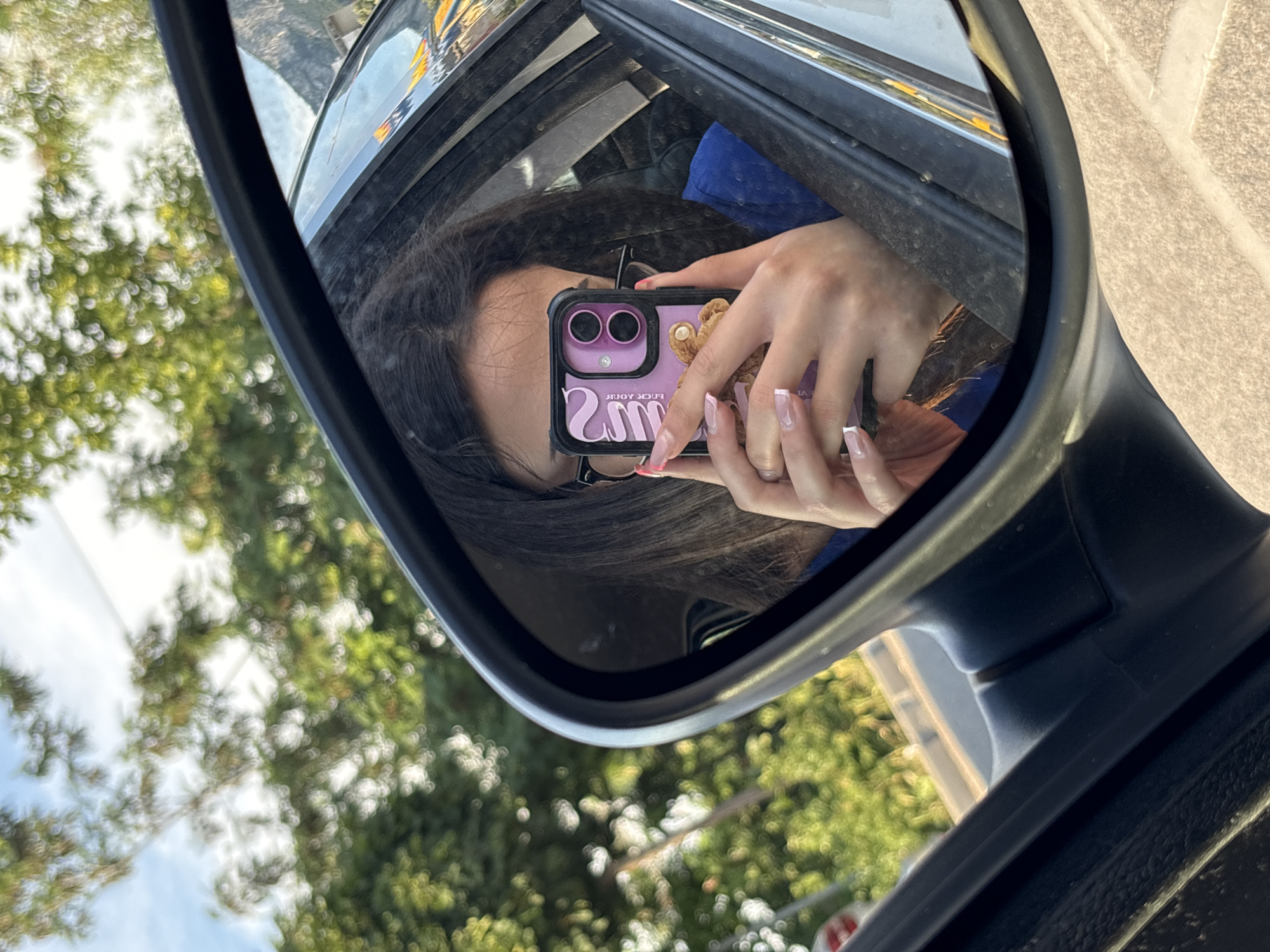 The image is a first-person perspective selfie taken in the side mirror of a car. The subject, a person with dark hair and glasses, is holding up a purple phone with a teddy bear case to take the photo. Their nails are painted with a French manicure. The background seen in the mirror and through the car window is an outdoor environment with trees and sky, suggesting a drive or a stop in a natural setting, possibly on the outskirts of Vratsa, Bulgaria, given the natural landscape. The side mirror frame is black, and the interior of the car is partially visible.
