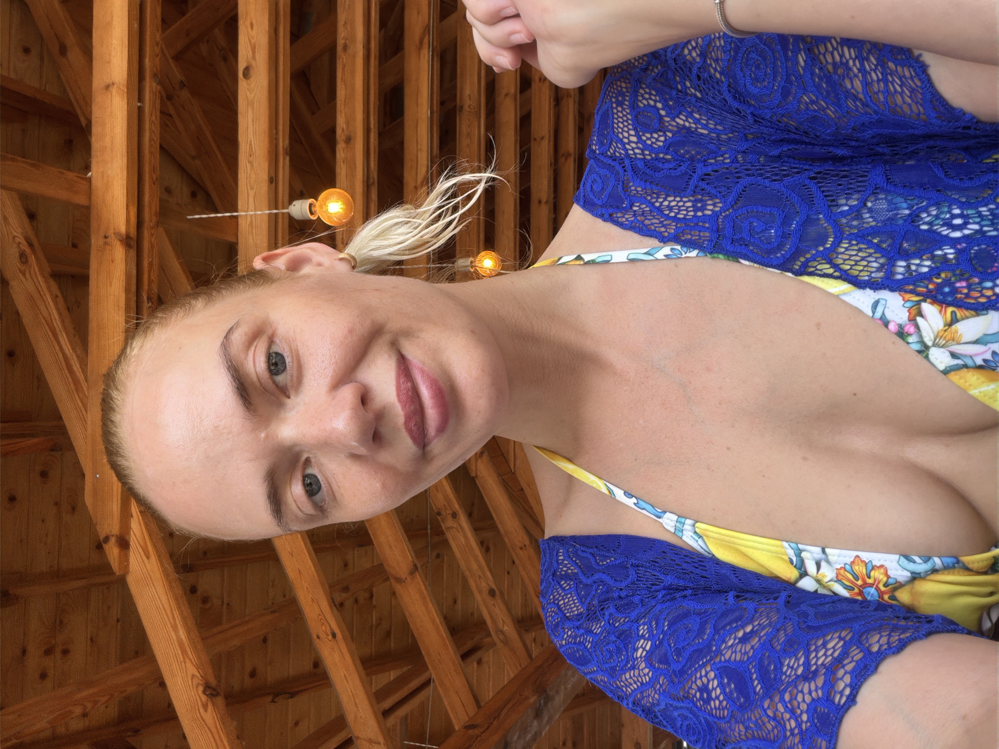 In an outdoor setting, a woman with blonde hair pulled back in a ponytail is captured in a close-up, selfie-style photograph. She is wearing a brightly patterned bikini top and a vibrant blue lace cover-up. Her head is slightly tilted, and she is looking towards the camera with a gentle smile. The background reveals a wooden structure, likely a roof with exposed beams and what appear to be decorative light fixtures hanging from the ceiling. The image has a warm, natural lighting. The location is identified as Puhoi, Moldova.