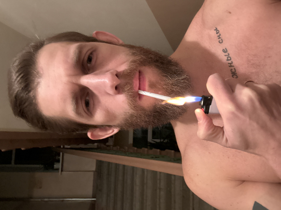 A bearded man is holding a cigarette in his mouth and using a lighter to light it. He is shirtless and has a tattoo on his upper chest. The background appears to be a room with a window overlooking an outdoor area. He is looking directly at the camera. The lighter has a flame with blue and yellow hues.