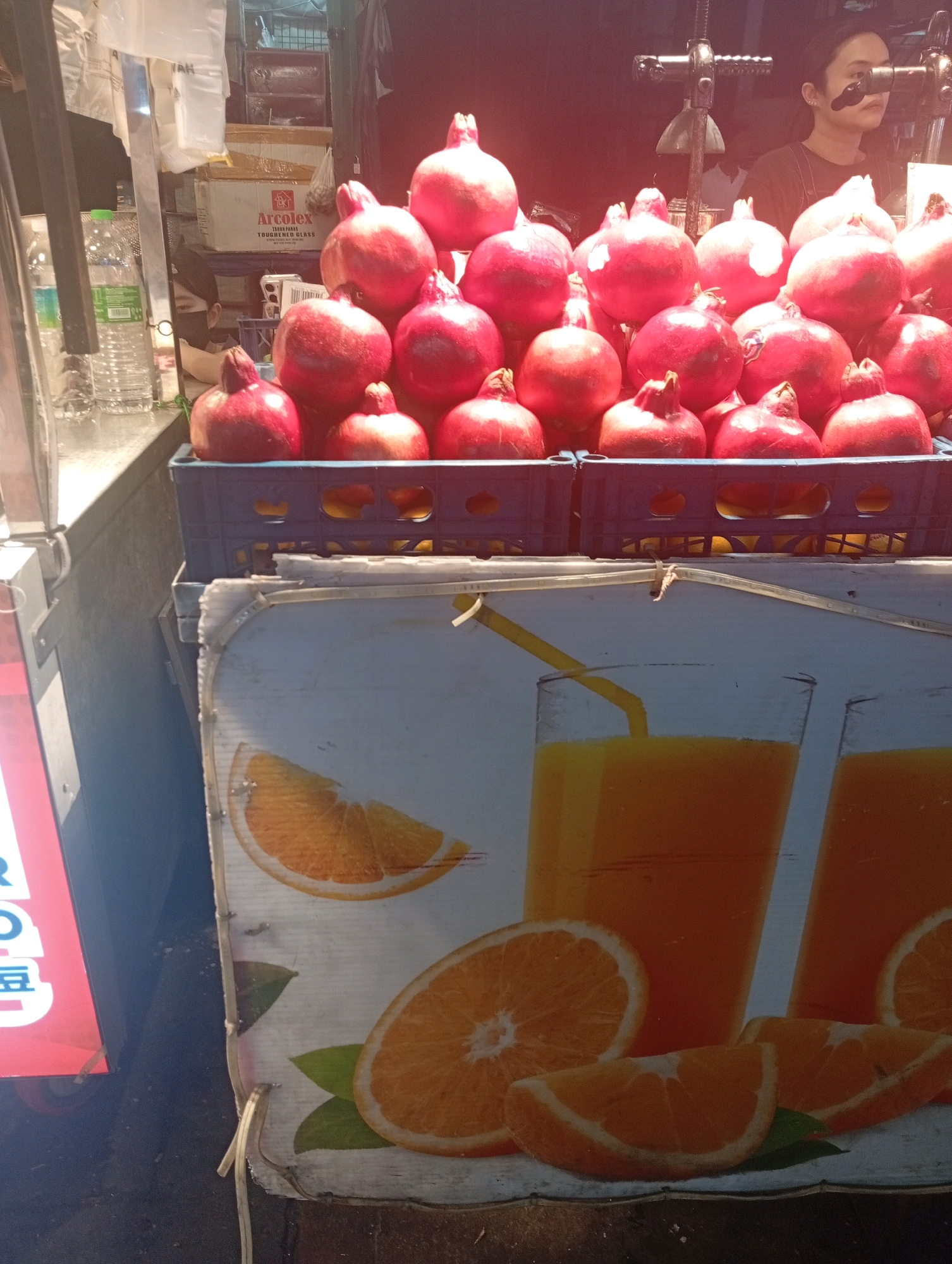 The image shows a street stall in Kuala Lumpur, Malaysia, with pomegranates and orange juice advertised. In the foreground, a sign depicts glasses of orange juice with orange slices. Behind the sign, several plastic crates are filled with ripe pomegranates. A woman is partially visible in the background, appearing to be working at the stall. Several bottles of water are also visible. The lighting suggests it is daytime, possibly late afternoon, with sunlight illuminating the fruit and the woman. The stall appears to be set up outdoors, typical of a bustling street market in Kuala Lumpur.
