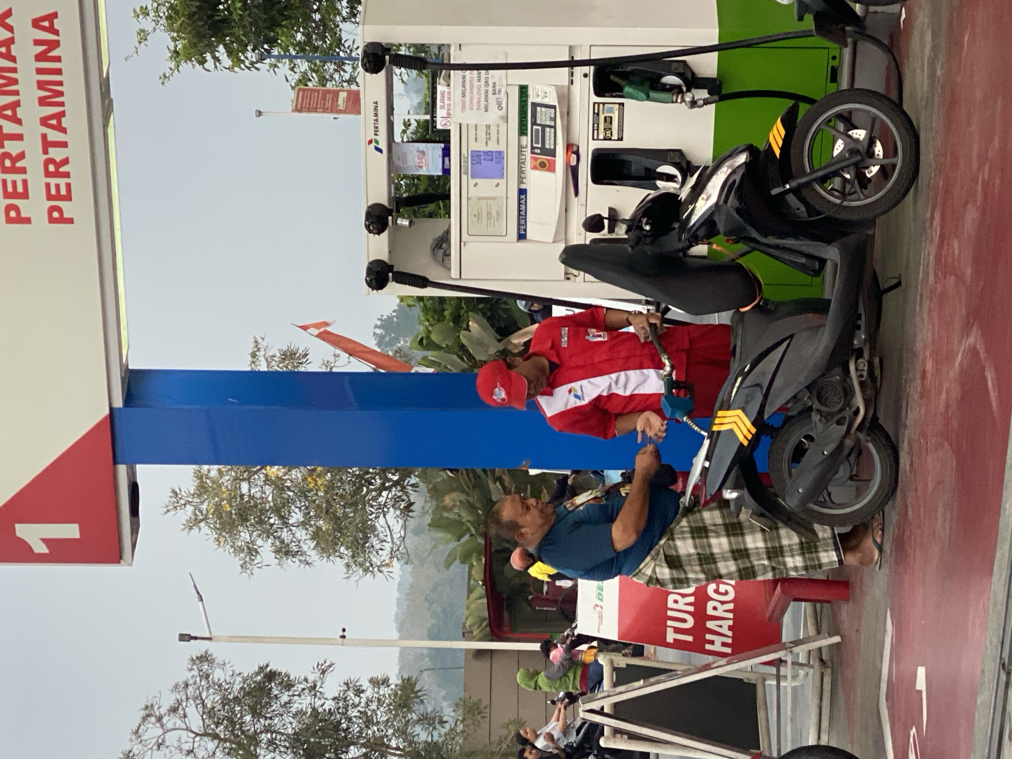 The image depicts a Pertamina gas station in Kasihan, Indonesia, where a man wearing a green and white checkered sarong is interacting with a gas station attendant in a red and white uniform. The man is handing cash to the attendant, who is holding a fuel nozzle. A black scooter is parked next to them, and a fuel hose is connected to it. The gas station has multiple fuel pumps, one of which displays prices for different fuel types like Pertamax and Pertalite. Other people and motorcycles are visible in the background, indicating a busy scene at the gas station. The environment suggests an outdoor setting, likely during daylight hours, with trees and a cloudy sky overhead.