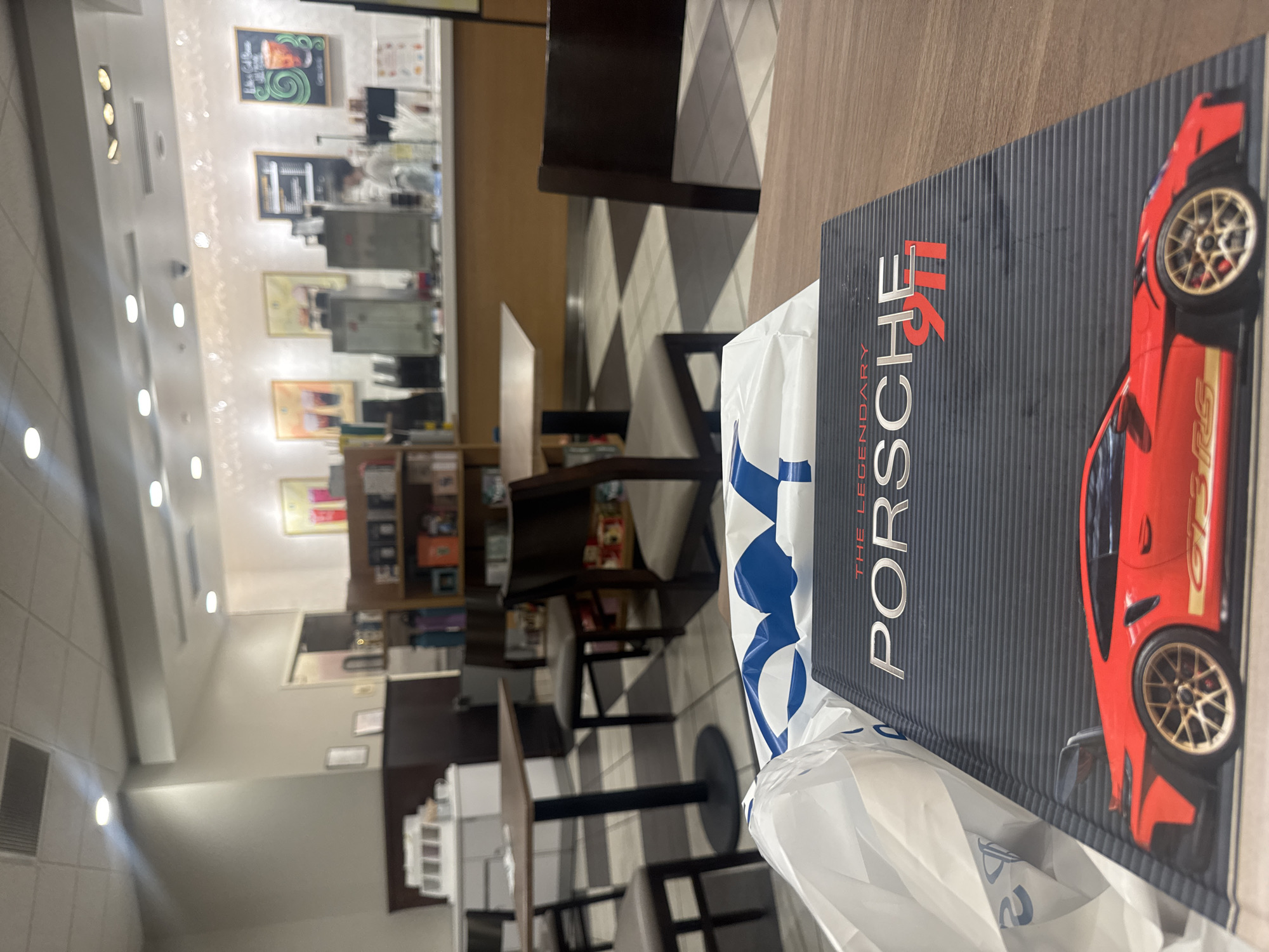 The image captures the interior of a cafe, strongly resembling a Barnes & Noble bookstore cafe, located in Boca Raton, Florida, United States of America. In the foreground, a large book titled "THE LEGENDARY PORSCHE 911" rests on a light brown wooden table. The book's cover features a detailed image of a red Porsche GT3 RS. A white plastic shopping bag with prominent blue markings partially covers the book and table to the left.
The midground reveals several empty tables with dark tops and matching chairs with light-colored upholstered seats, arranged on a checkered floor composed of dark and light tiles. A wooden bookshelf stocked with various items is visible on the right side of the frame.
The background features a long service counter where an individual, likely an employee, stands behind an assortment of beverage machines and preparation equipment. Above the counter, the wall displays several framed pictures or menu boards, including one with a green swirled design and text. The entire space is illuminated by numerous ceiling-mounted lights, and the walls are a uniform light beige color. The setting appears clean and orderly, indicative of a commercial establishment.
