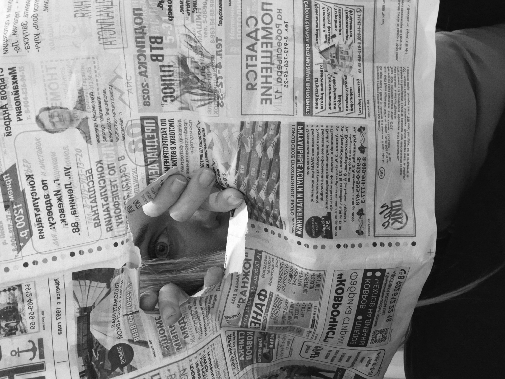 A person is looking through a torn newspaper with Russian language text and advertisements. The newspaper advertises items such as carpets, real estate, and home appliances. One advertisement mentions the city of Votkinsk.