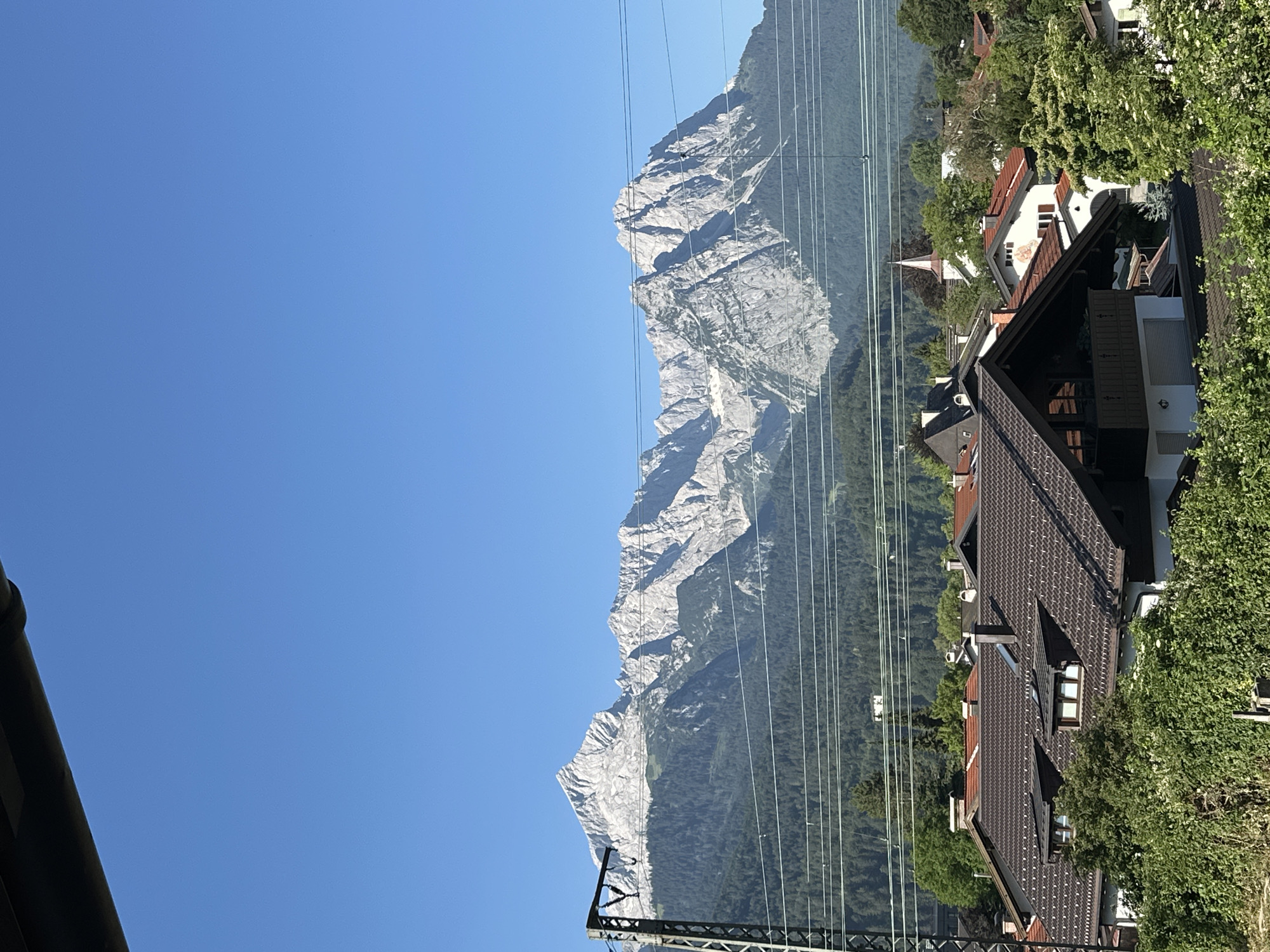 The image presents a serene daytime view of a suburban area in Garmisch-Partenkirchen, Germany, with a dramatic mountain backdrop. In the foreground, several tiled roofs of residential houses are visible, interspersed with lush green foliage. Some houses feature traditional architectural elements such as dormer windows and balconies.
Beyond the houses, a dense forest stretches across the middle ground, creating a transition zone to the imposing mountains that dominate the background. These mountains are characterized by their rugged, rocky peaks, showcasing a stark contrast between the natural landscape and the human settlement. The sky above is a clear, bright blue, suggesting fair weather. Electrical wires and a power line tower are also present, indicating the presence of infrastructure. A small church steeple is visible peeking out from behind the trees in the distance.