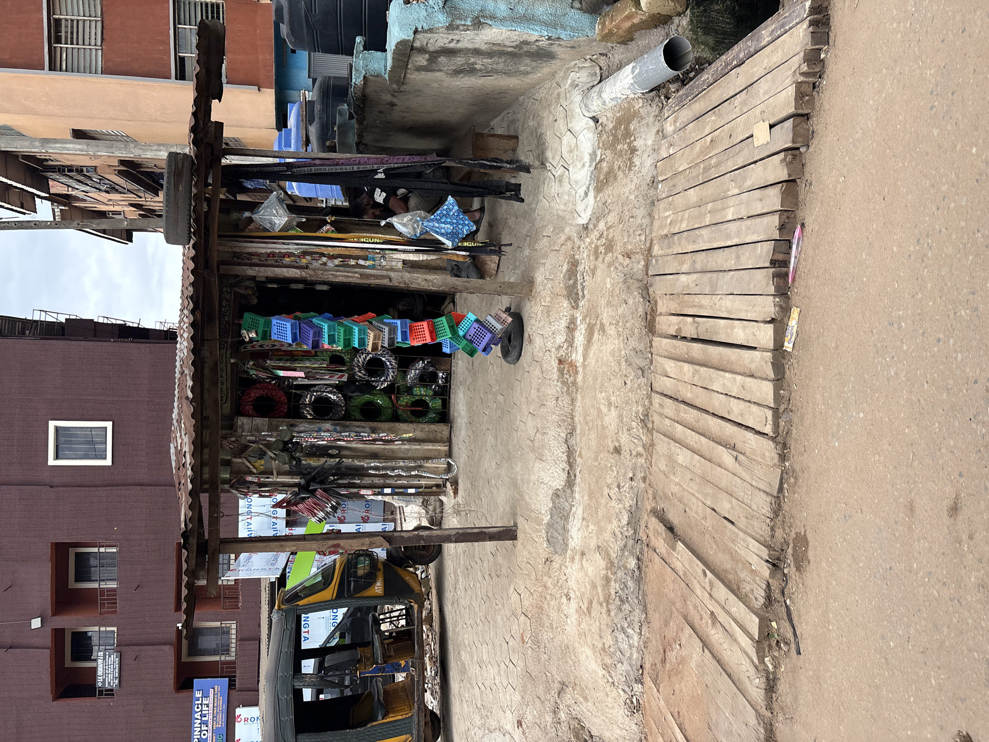 This is an outdoor medium shot of a roadside stall and its surroundings in Nkpor, Nigeria, on a cloudy day. The stall has a corrugated metal roof and displays various items for sale, including colorful plastic baskets, coiled wires, and other miscellaneous goods. A person is seated inside the stall on a wooden bench, partially obscured by hanging merchandise. To the left of the stall, a yellow and black auto-rickshaw is parked on a dirt and paved surface. In the background, multi-story buildings with balconies and windows are visible. A concrete wall runs along the right side of the scene, topped with large black water tanks. A grey drainage pipe extends from the wall towards the foreground. A wooden walkway, constructed from planks, is laid over the ground in front of the stall. The ground surface is a mix of dirt, concrete pavers with a hexagonal pattern, and the wooden walkway. Scattered on the ground are some discarded wrappers and a small black object.