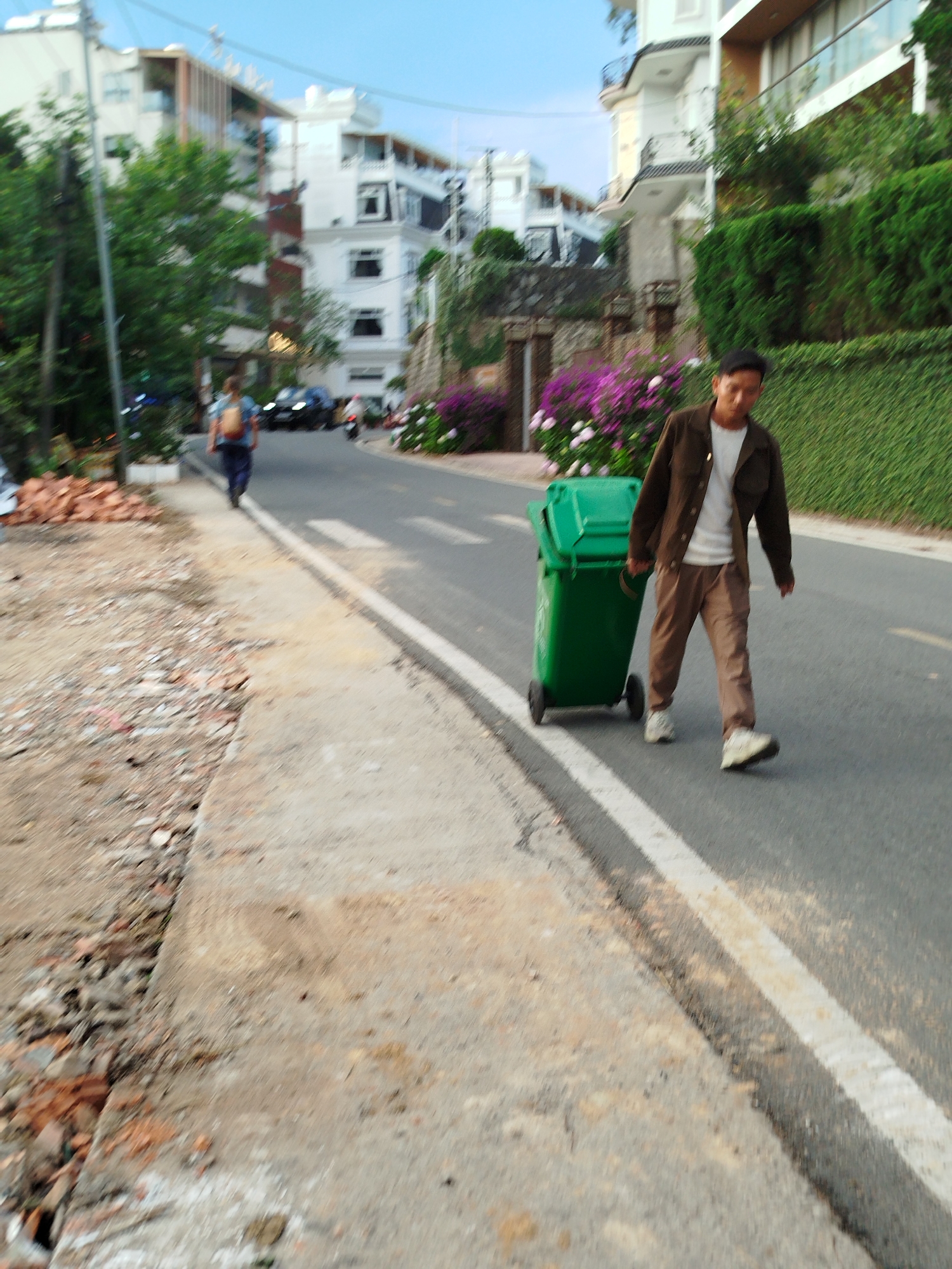 A road scene in Da Lat, Vietnam, features a man pulling a green rolling trash bin along a road marked with a white line. The man is wearing tan trousers, a white t-shirt, and a brown jacket. Behind him, another person walks down the road, away from the viewer. Buildings and green vegetation border the road, including flowering shrubs. The side of the road closest to the viewer shows construction debris.