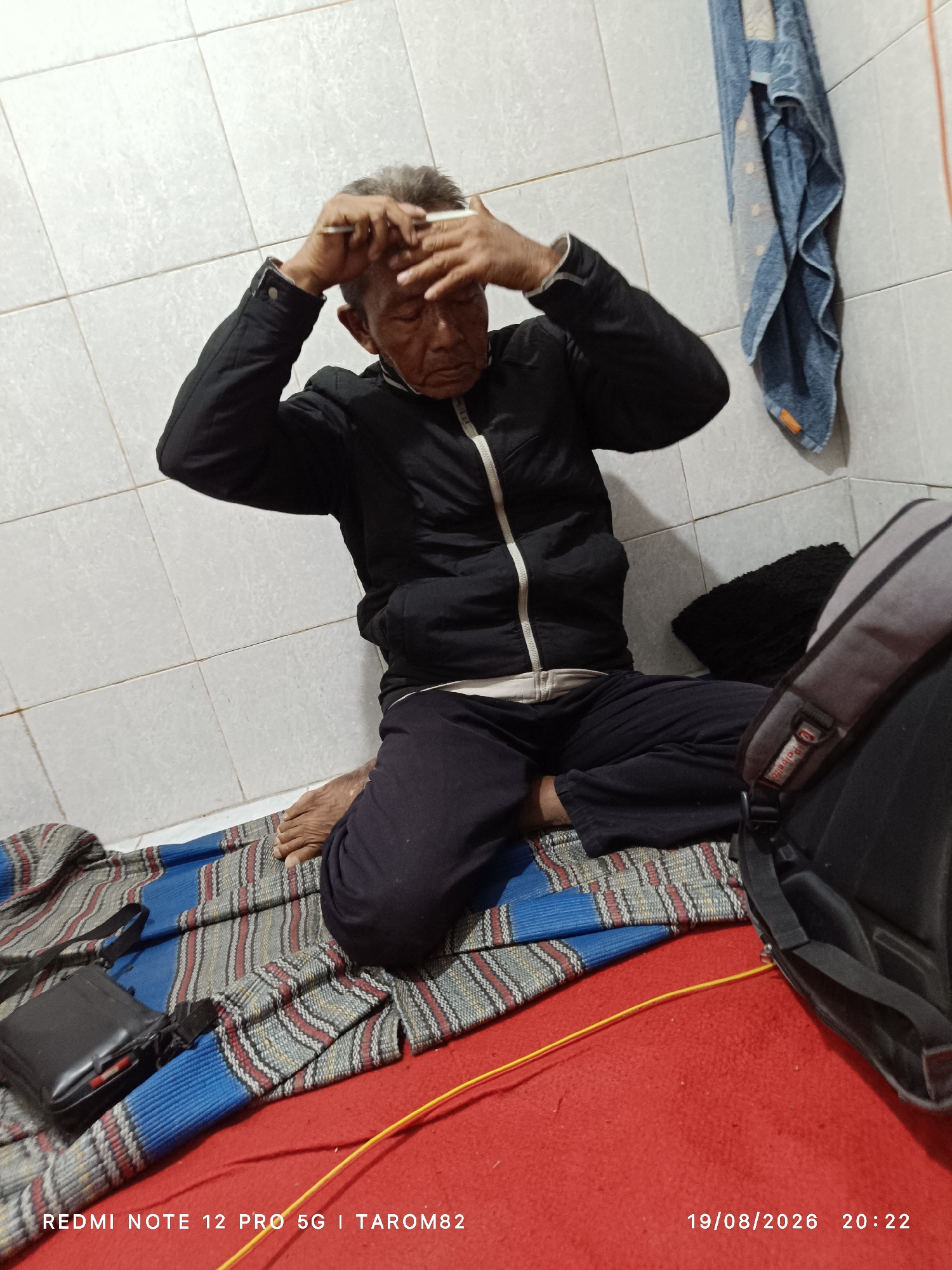 An elderly East Asian male is seated on a striped mat on a red floor in what appears to be a bathroom, given the tiled walls and a blue towel hanging on the wall. He is wearing a black puffer jacket and dark pants. His hands are raised to his head, and he appears to be combing his hair with a thin object, possibly a comb or a pencil. To his left, a black leather bag is placed on the mat. To his right, a portion of a grey backpack is visible. The bottom right corner of the image displays a timestamp: "19/08/2026 20:22" and the text "REDMI NOTE 12 PRO 5G | TAROM82". The environment is dimly lit, suggesting the photo was taken indoors. The overall scene is mundane and domestic. The specific location within Sidodadi, Indonesia, cannot be determined from the visual information alone.