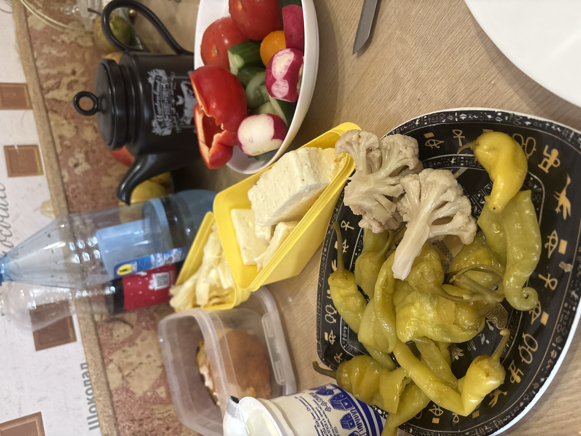 The image shows a table with various food items. There are sliced tomatoes, cucumbers, radishes, and a red bell pepper in a white bowl. There is also a yellow container with chunks of white cheese. A black teapot sits beside the bowl. A plate with pickled yellow peppers and a few pieces of cauliflower is also present. Other items include a plastic water bottle, a container with food inside, and a container of yogurt. The table has a patterned tablecloth with what appears to be Cyrillic text including the word "Шоколад" (Chocolate). The text on the yogurt container indicates it's a dairy product. The scene appears to be indoors. The image does not contain enough information to determine the location in Krasnoyarsk, Russia.