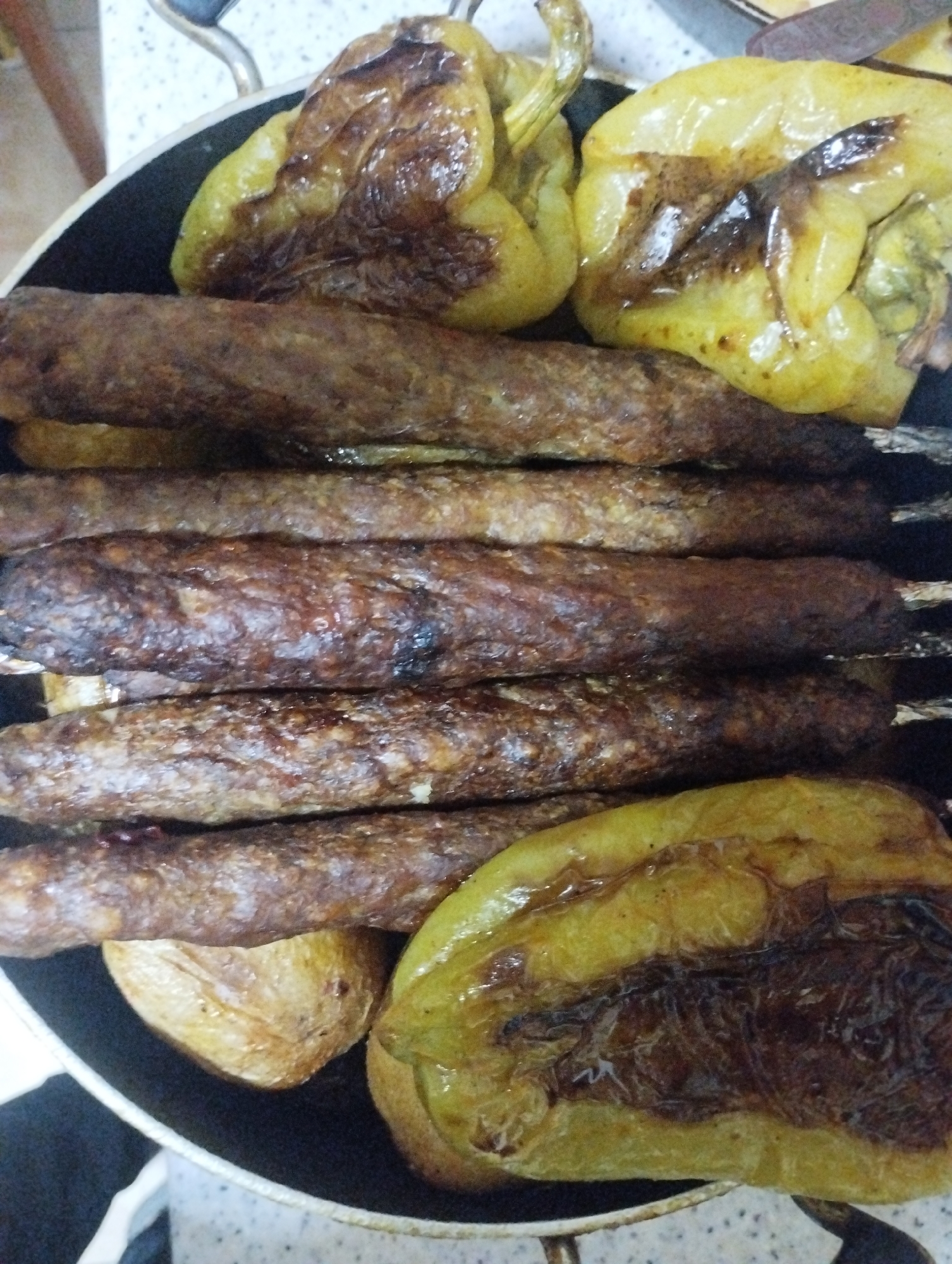A close-up, overhead shot shows a black frying pan containing a meal of grilled sausages and roasted bell peppers. The sausages are arranged horizontally in the center of the pan, appearing brown and slightly charred. Above and below the sausages are pieces of roasted yellow bell peppers, some showing signs of charring and browning. Two whole roasted potatoes are visible at the bottom of the pan. The setting appears to be a kitchen, with a white countertop and a glimpse of kitchenware in the background. This meal was prepared in the city of Bilajari, Azerbaijan.