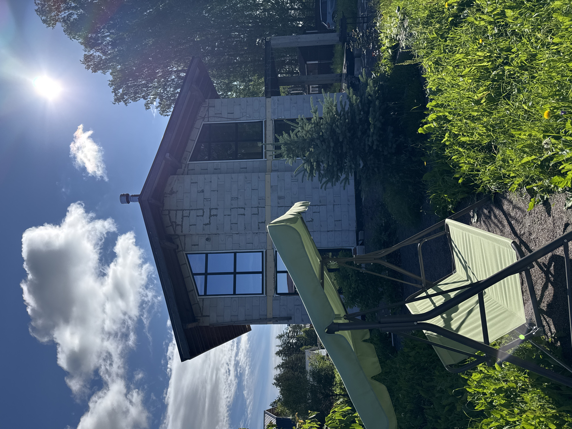 The image depicts a partially constructed house in Steklyannyi, Russia, on a sunny day. The house is made of grey cinder blocks and has large, dark-paned windows. A dark-colored pitched roof with visible wooden rafters is installed. A chimney pipe protrudes from the roof. In the foreground, a green garden swing with a matching canopy is positioned on a gravel path. Lush green grass and foliage surround the swing and the house. Tall trees, including what appears to be a fir tree near the house, are visible. The sky is blue with scattered white clouds, and the sun is shining brightly, causing lens flare. A dark-colored vehicle and a small shed-like structure are visible in the background. The scene suggests a residential area undergoing construction.