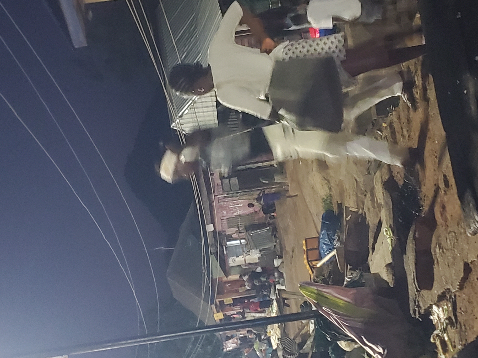 The image depicts a dimly lit outdoor scene, possibly a market or a street in Gwong, Nigeria. Several people are present, some blurred due to motion, suggesting movement or low light conditions. In the foreground, two individuals are prominent. One, seen from the back, wears a white t-shirt and a patterned skirt, holding a dark bag. Another person, to their left and also blurred, appears to be wearing white pants and a dark top.
The environment consists of dirt ground with scattered debris and what seem to be structures with corrugated metal roofs in the background. There are also utility wires visible overhead. Some goods, possibly produce in crates and wrapped items, are visible on tables or stands, indicating a commercial setting. The overall atmosphere is one of a bustling evening in a community with limited resources.
