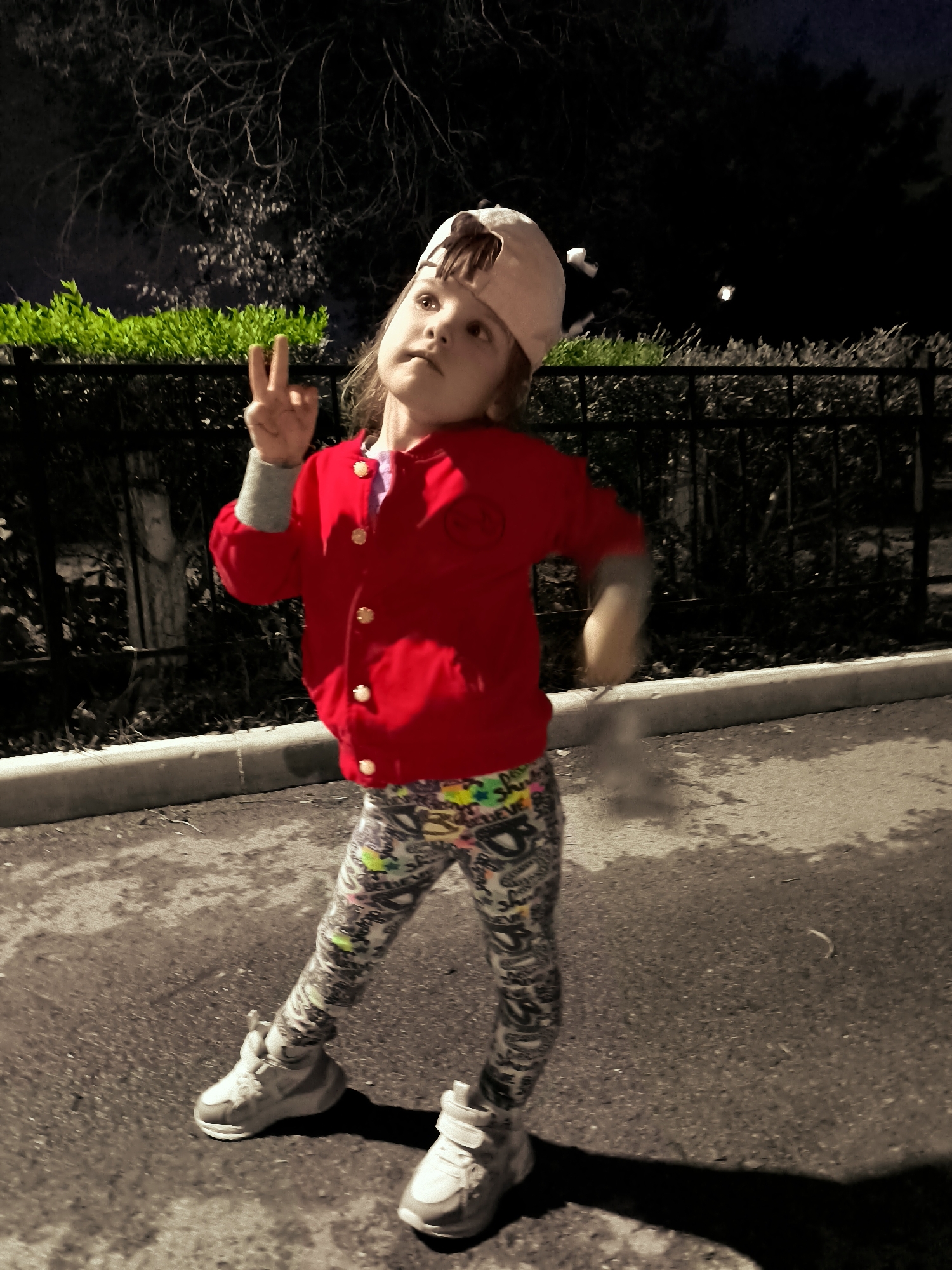 A young girl wearing a red jacket, patterned leggings, and a light-colored baseball cap stands on a paved surface, possibly a sidewalk or parking lot, at night. She is holding up two fingers on her left hand, giving a peace sign. The background features a dark, shadowy area with trees and a black metal fence, as well as some green bushes. The lighting suggests artificial illumination. The location is identified as the city administration area of Qarağandı, Kazakhstan.