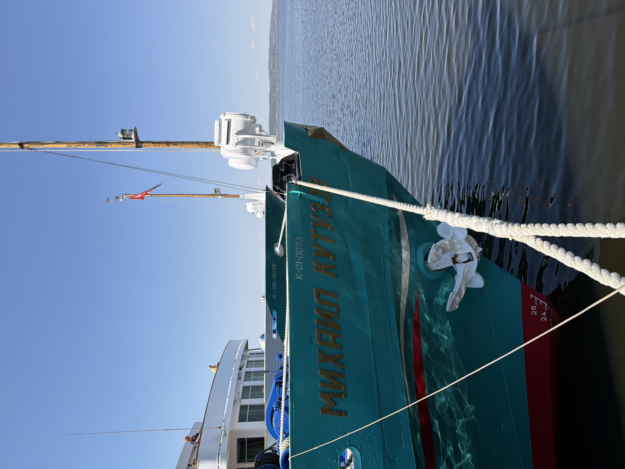 Here's a detailed description of the image:
The image depicts a close-up view of the side of a river cruise ship named "Mikhail Kutuzov," a project 305 type ship. The ship is painted in blue-green with red and white accents. The name "МИХАИЛ КУТУЗОВ" is written in gold Cyrillic letters on its side. Also on the hull are the markings "K-06-0035" and "K-01-0033."
A thick white rope secures the vessel to the dock. A white anchor is visible near the bow. Draft marks are painted on the hull.
The water alongside the ship is relatively calm, with some slight ripples.
Part of the upper deck and superstructure of the ship is visible. The sky above is a clear, light blue. In the background, there is a view of a body of water.
The image was likely taken in the city of Chaikovsky, Russia, where similar river cruise ships operate on the Kama River. The orientation of the photo is vertical and the image has been rotated.
