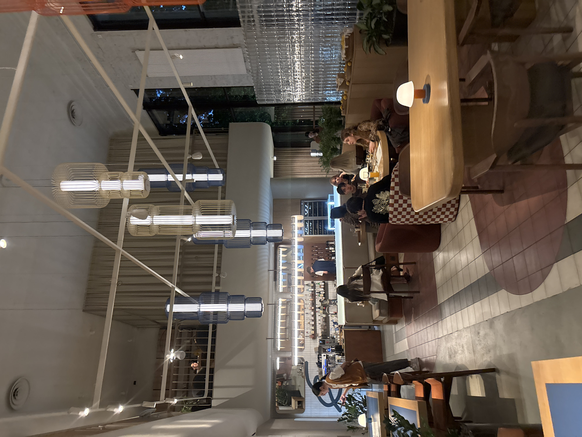 The image shows the interior of a restaurant in Novaya Usman', Russia. The restaurant has a modern design with high ceilings and large windows. There are several dining areas, including a bar area, a lounge area, and a main dining area. The restaurant is dimly lit with pendant lights and table lamps. There are several patrons dining and socializing in the restaurant. Some are seated at tables, while others are standing at the bar. A server is visible in the bar area, and a bartender is behind the bar. The overall atmosphere of the restaurant appears to be relaxed and sophisticated.