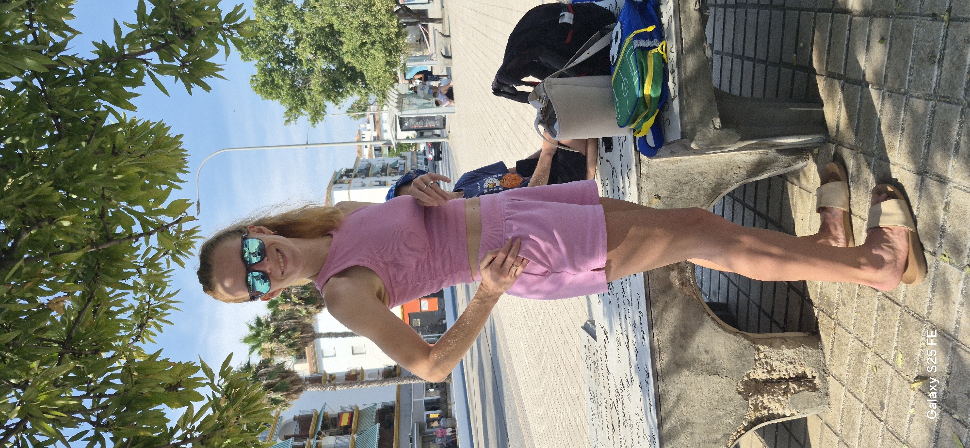 A young woman with blonde hair and sunglasses is standing on a sidewalk in what appears to be a sunny outdoor setting. She is wearing a pink sleeveless crop top and matching pink shorts. Her left hand is on her hip, and her right hand is resting on her right thigh. She is smiling.
In the background, there are trees with green leaves, a street light, buildings, and people sitting at what looks like a bus stop. There are also backpacks and a purse on a bench next to her. The ground is paved with light-colored bricks and has shadows cast on it. The sky is clear and blue.
The image is oriented sideways, so the woman is standing upright when viewed rotated. A watermark at the bottom right corner reads "Galaxy S25 FE". The overall impression is of a casual, bright day, likely in a tourist or urban area.