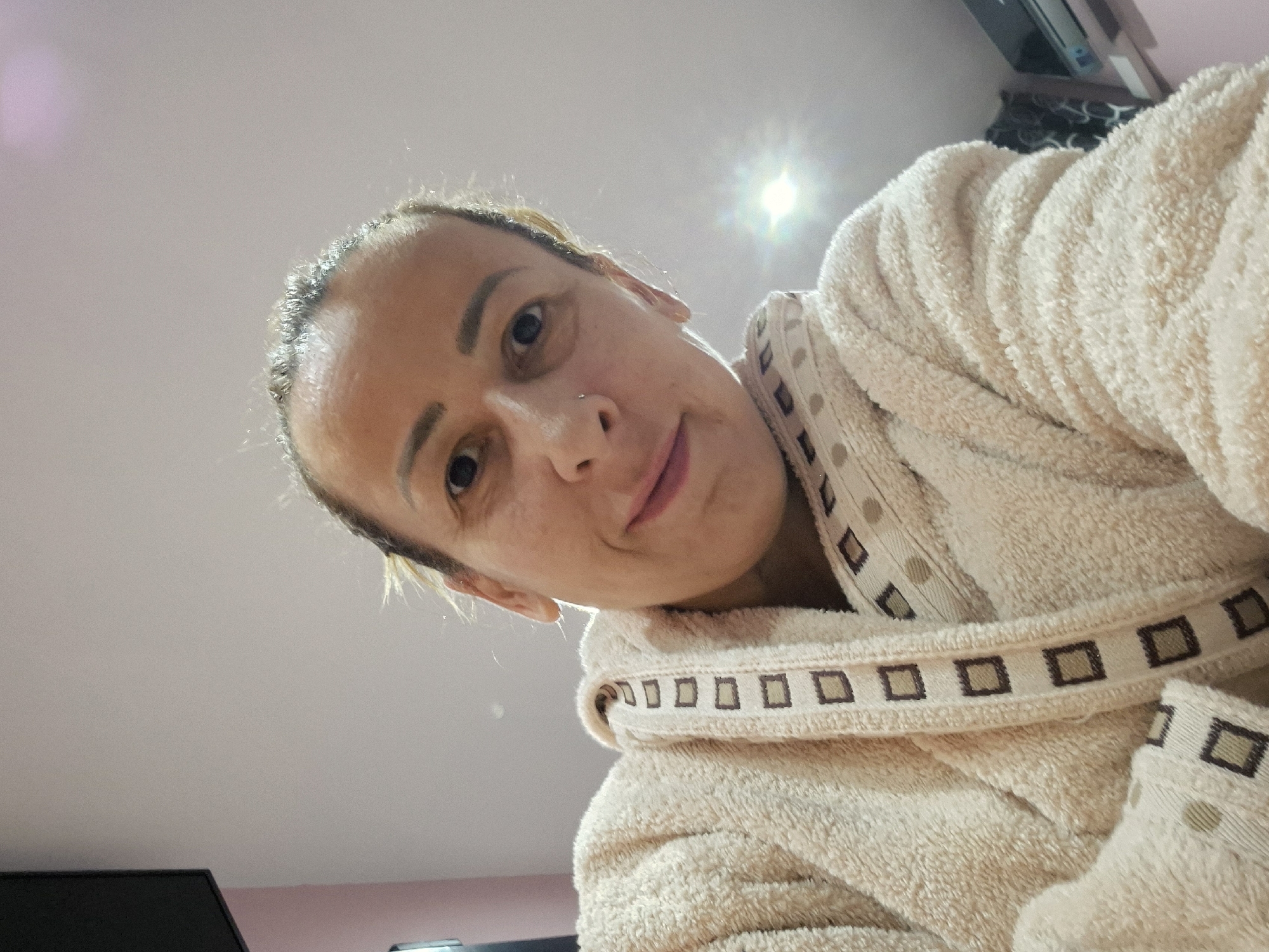 A woman is angled downward, presumably taking a selfie. She has fair skin, brown eyes, a nose piercing, and is wearing a beige terrycloth bathrobe with a decorative pattern of squares and dots on the collar and cuffs. The background is a pale pink wall and a portion of a black television. The location is an indoor space, possibly a bedroom or living room, in an unknown city.