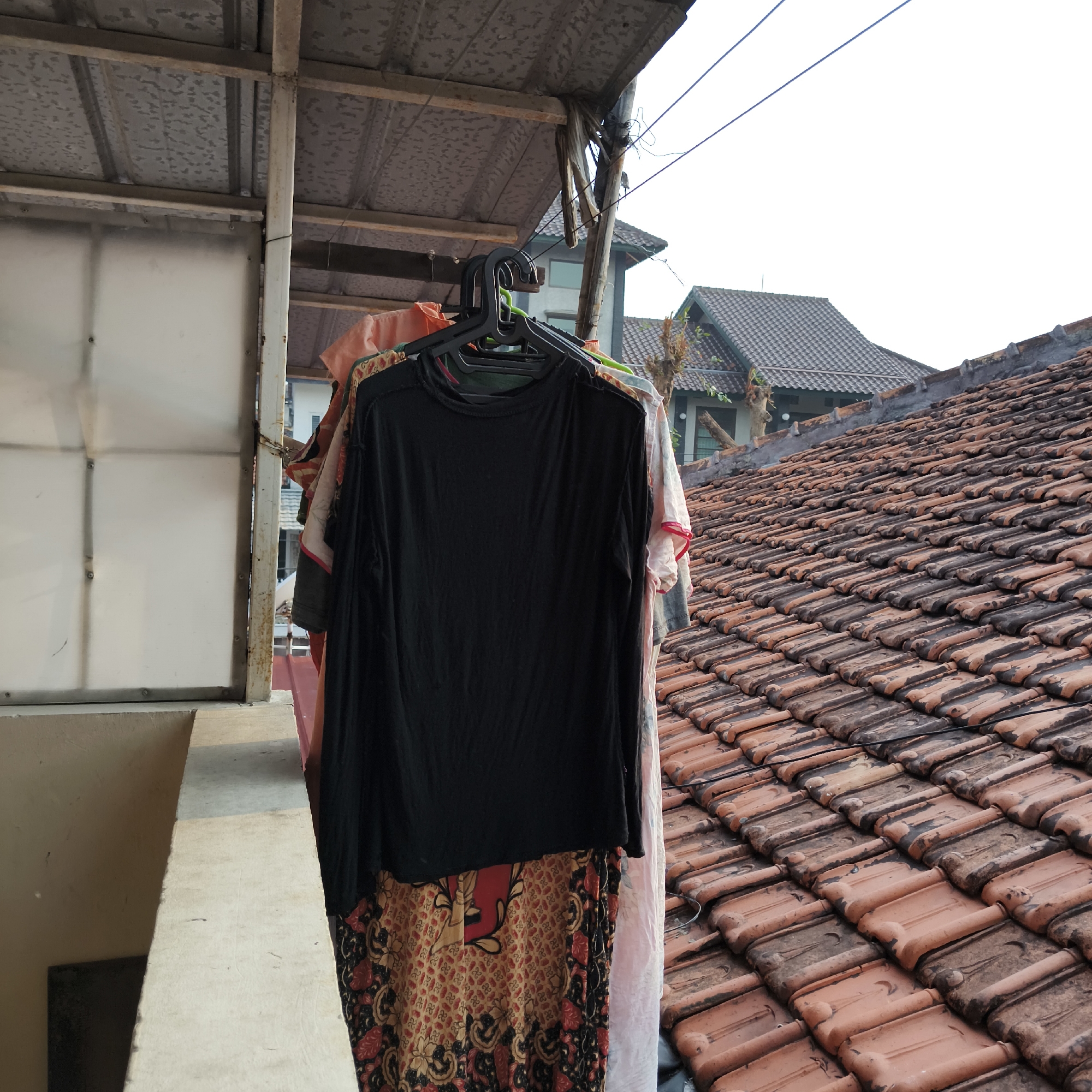 The image shows a close-up of several pieces of clothing hanging on black plastic hangers. The most prominent item is a plain black long-sleeved shirt. Beneath it, a patterned fabric, possibly a sarong or skirt, with red, yellow, and black designs is visible. Other garments, including a pinkish garment and a white garment with red trim, are also hanging. The clothing is suspended from a clothesline system attached to an overhanging structure with a corrugated metal roof. In the background, a tiled roof of a building is visible, with part of another building and a clear sky also in view. The scene suggests an outdoor drying area for laundry. The overall impression is a typical residential setting in South Jakarta, Indonesia, likely on a rooftop or balcony.