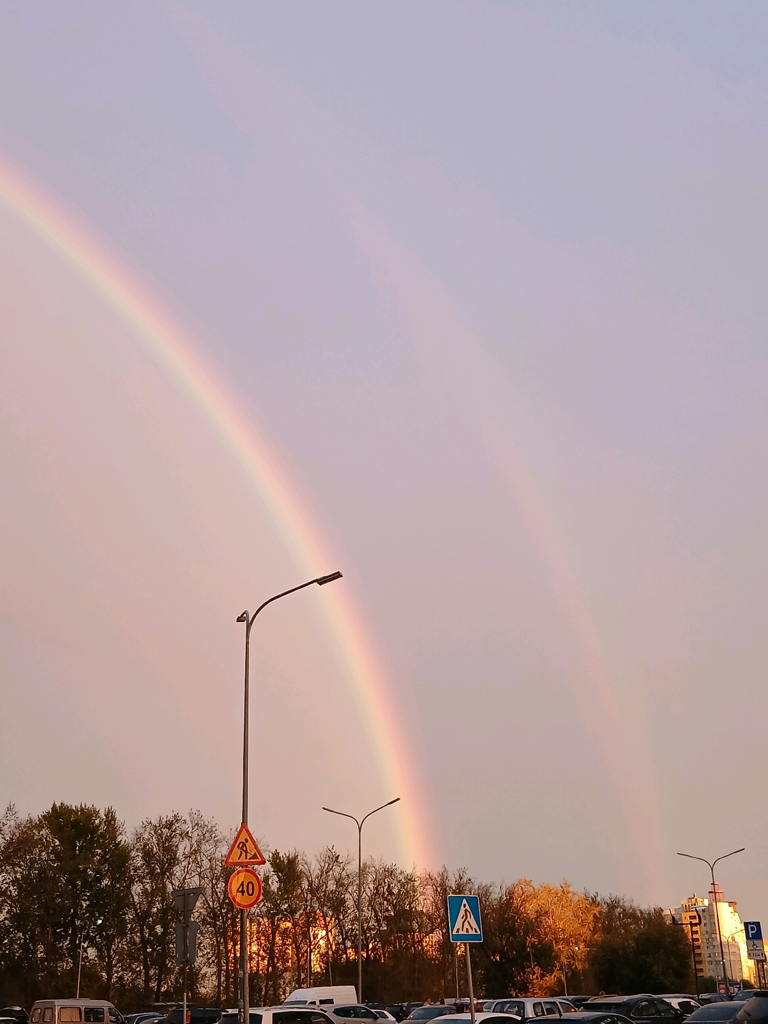 A double rainbow arcs across a pale lavender sky over a parking lot in Nizhny Novgorod, Russia. The sky is filled with pastel hues, with a faint pinkish glow on the horizon. A single streetlamp with a curved pole stands in the foreground, its light emitting from the right side. Below, a line of trees with autumn foliage forms a dense backdrop. Various traffic signs are visible, including a yellow triangular sign depicting a construction worker and a red circular sign with the number "40," indicating a speed limit. A blue square sign with a white pedestrian symbol is also present. In the distance, a modern apartment building with several floors stands out against the trees. Cars are parked in rows, and a white van is parked on the left side of the frame. The scene appears to be captured during sunset, as evidenced by the warm light illuminating the trees and the soft colors in the sky.