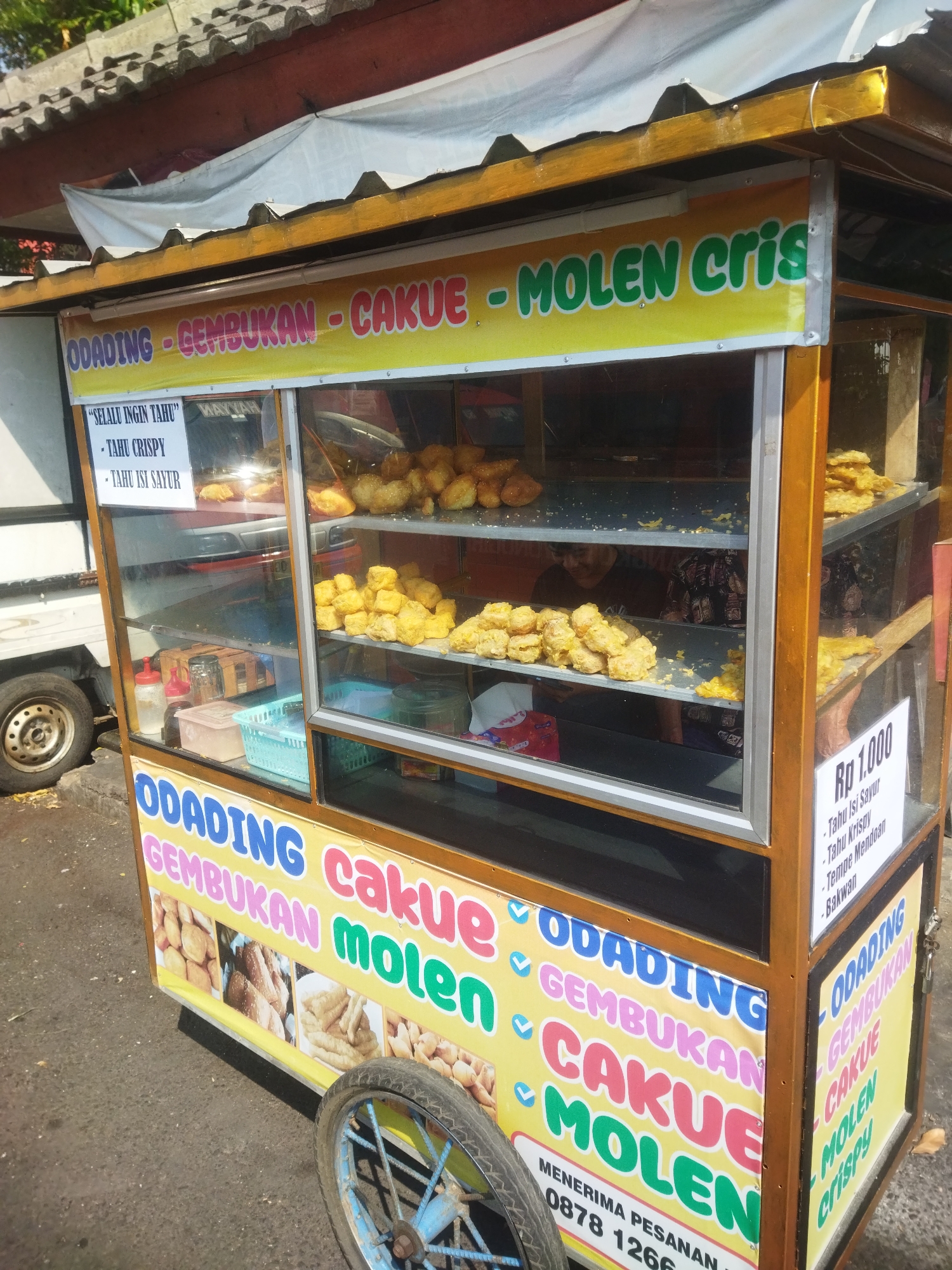 A street food cart is pictured in the city of Wonogiri, Indonesia. The cart is adorned with colorful banners advertising food items like "ODADING," "GEMBUKAN," "CAKUE," and "MOLEN CRISPY." One banner, located on the front of the cart, displays images of fried snacks. Another banner on the side lists prices, including "Rp 1.000" for "Tahu Isi Sayur," "Tahu Krispy," and "Tempe Mendoan." Inside the cart, glass shelves display fried foods, appearing to be a variety of fritters and fried tofu. A handwritten sign on the inside glass reads, "SELALU INGIN TAHU" (Always Want Tofu), followed by "TAHU CRISPY" and "TAHU ISI SAYUR" (Tofu with Vegetables). The cart is equipped with a wheel, suggesting it is mobile. The surrounding environment is an outdoor setting with a paved surface, indicating a street or roadside location. A portion of a white vehicle is visible to the left of the cart. The roof of a building with tiled roofing is visible above the cart.