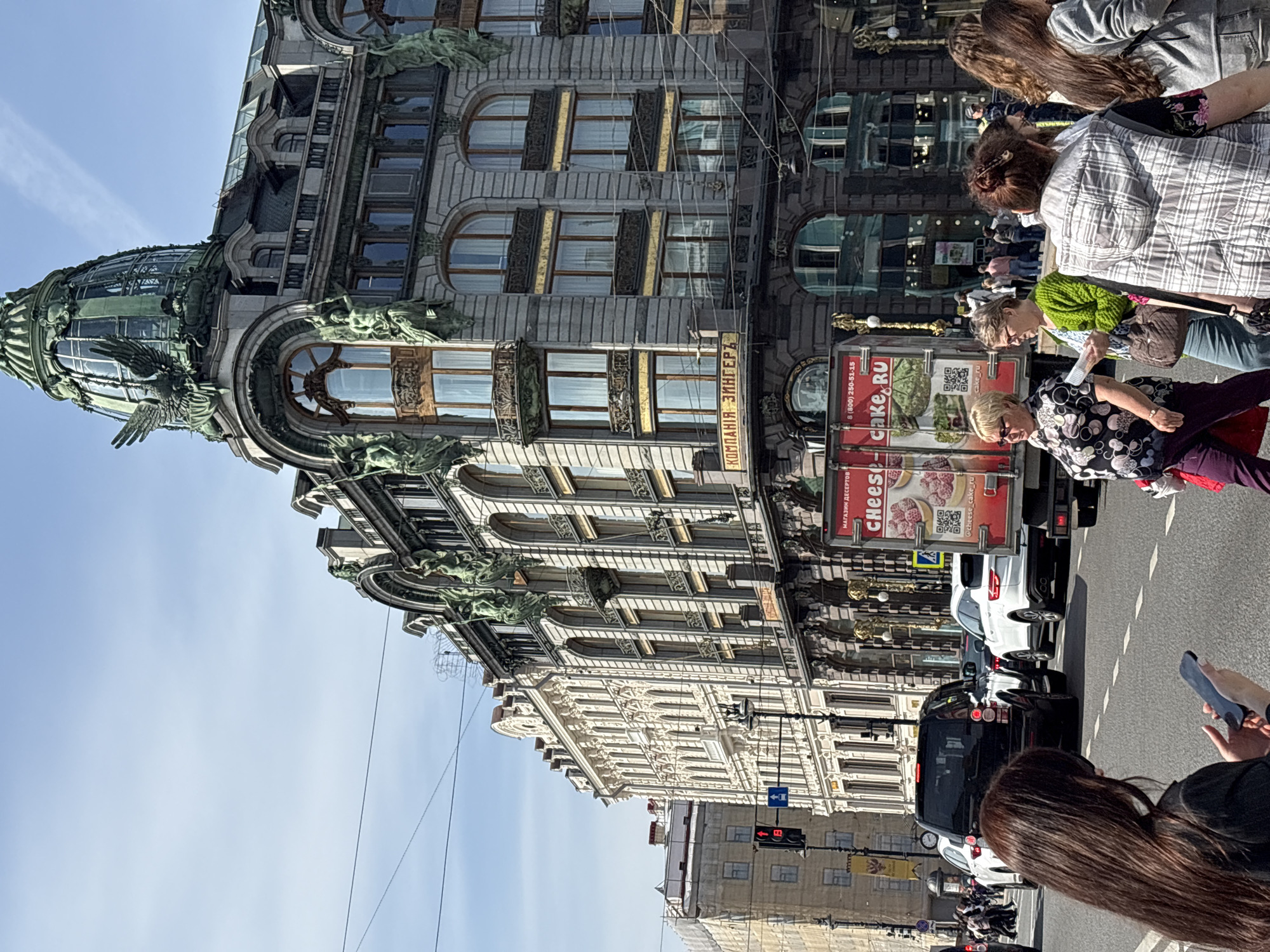 The image shows the Singer House, also known as the Dom Knigi, a prominent building located at Nevsky Prospekt 28 in Saint Petersburg, Russia. The architectural style is Art Nouveau, characterized by its ornate facade, large windows, and the distinctive glass dome topped with a sculptural eagle. People are visible on the sidewalk, and traffic is present on the street in front of the building, including cars and a truck bearing advertising for a dessert shop. The sky is clear and blue.