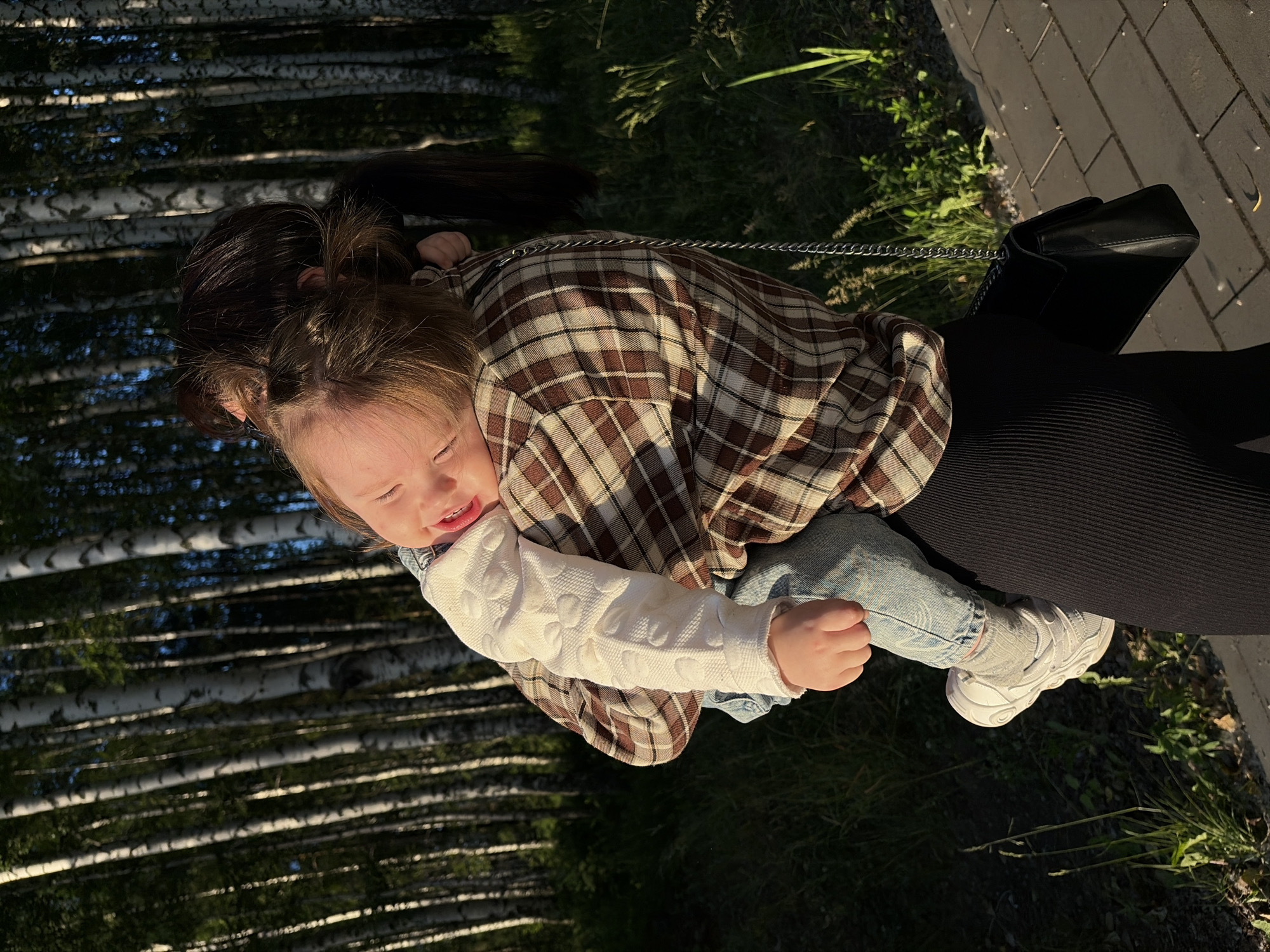 A person, likely a parent, is holding a young child in their arms. The child is facing away from the camera and appears to be happy, with their mouth open and eyes closed. They are wearing a white long-sleeved shirt with a patterned texture, blue jeans, and white sneakers. The person holding the child is wearing a brown and beige plaid shirt and black ribbed pants. A black purse with a silver chain strap is slung over their shoulder.
The background consists of a dense forest of birch trees, with their white trunks and dark foliage visible. Sunlight is filtering through the trees, creating dappled light on the ground and illuminating the scene. There is also some green grass and vegetation in the foreground, and a paved walkway, possibly in a park or public area. The overall setting suggests an outdoor activity during the day, possibly in the late afternoon or early evening due to the warm, golden light.
Considering the context of Chaikovsky, Russia, this scene could be taking place in one of the city's numerous parks or recreational areas, many of which are characterized by abundant birch forests and walking paths, especially near the Kama River. The presence of a paved walkway and manicured green spaces further supports the idea of a public park or a well-maintained outdoor area within or on the outskirts of the city.