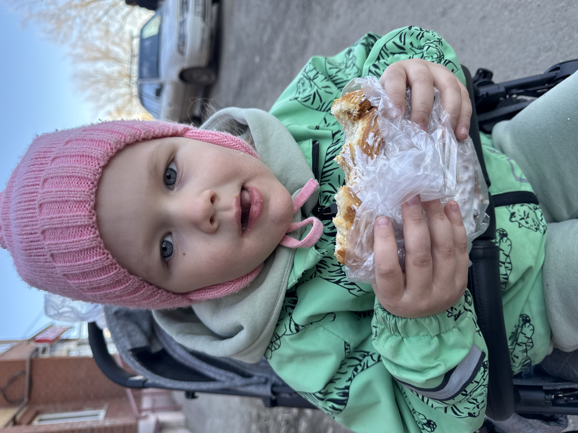 A young child, presumed to be a girl, is seated in a stroller outdoors. She wears a pink knit hat and a light green jacket with black tiger designs. She holds a sweet pastry wrapped in a clear plastic bag in her right hand, indicating she has been eating it. Her facial expression suggests she may be about to say something. The background shows an urban street scene. A light-colored car is partially visible in the background. The location is Ферма, Russia.