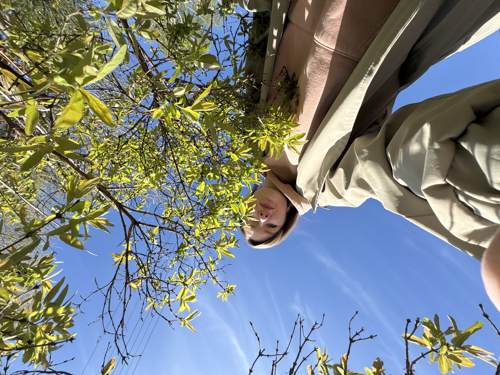 A low-angle shot shows a person and a tree against a clear blue sky. The person, likely female, appears upside down, with her face in the upper center of the frame. She has fair skin and light brown hair, and is wearing a light brown jacket under a tan-colored coat. The tree is on the left, its branches framing the blue sky, with green and yellow leaves. The photo is likely taken outdoors in a sunny environment. There is no clear indication of Italmas, Russia, in the image.
