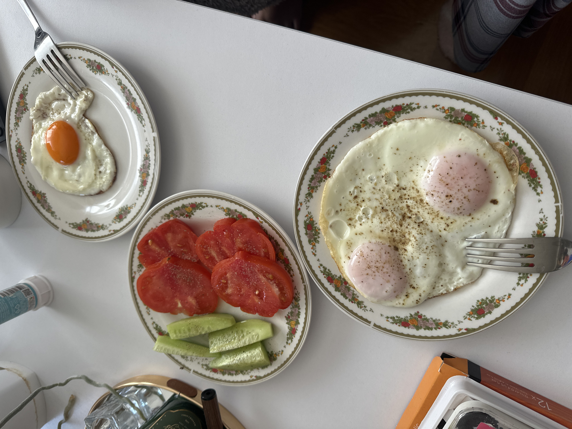 The image depicts a breakfast scene. Two plates contain fried eggs, one with a single egg and the other with two eggs, seasoned with black pepper. A third plate features sliced tomatoes and cucumbers. Silverware, specifically forks, are present on the egg plates. The breakfast is set on a white surface. Objects such as a bottle, a box, and a decorative item are visible in the lower left and right corners of the image. The location is not explicitly identifiable as Budva, Montenegro, based on the provided visual information alone. There is no evidence of people, specific actions, or interactions in the image.