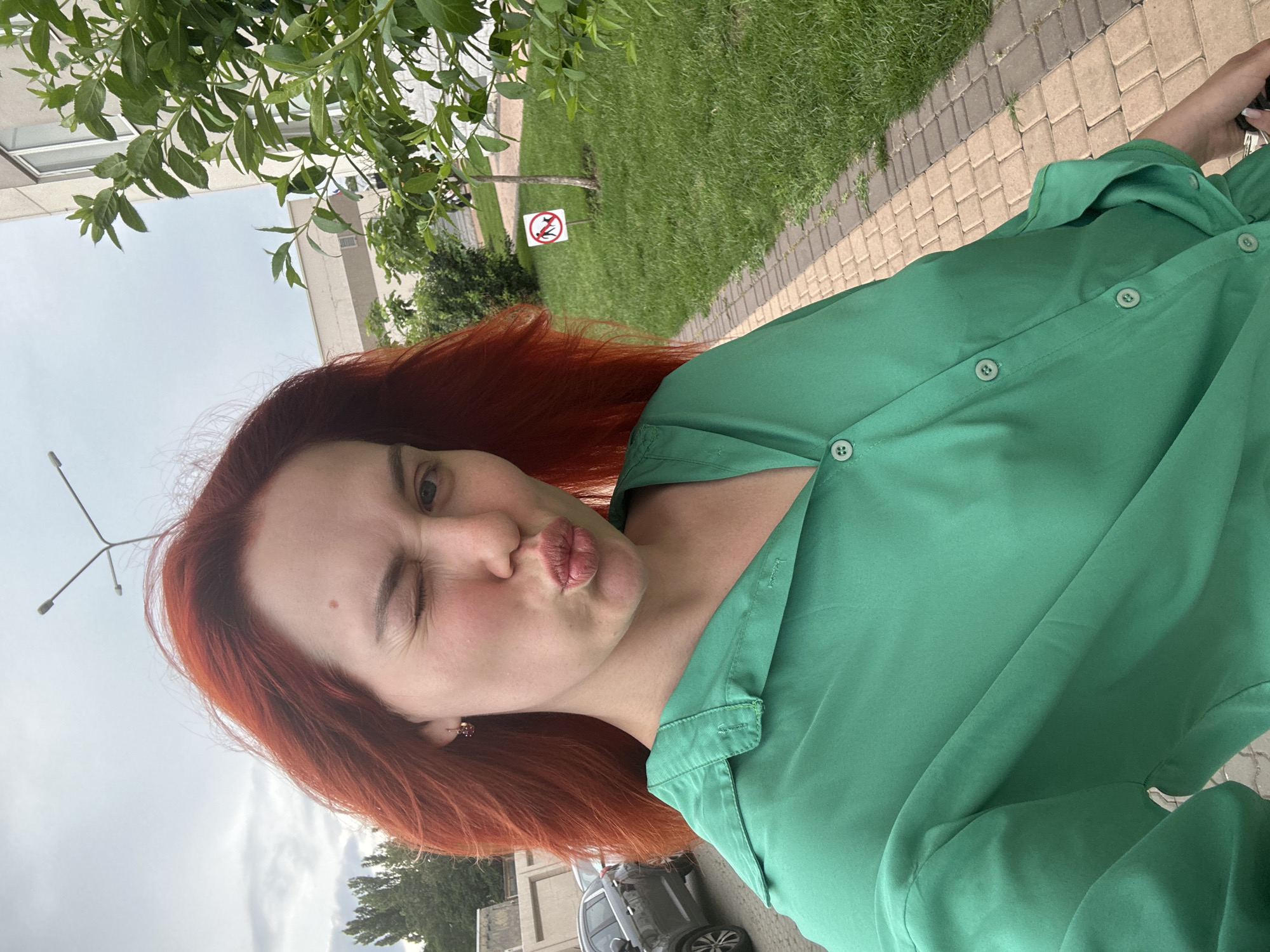 A young woman with bright red hair is taking a selfie outdoors, winking and puckering her lips. She is wearing a bright green button-down shirt. In the background, there is a paved walkway, a grassy area with a "no dogs" sign, trees, a building, and a cloudy sky. The lighting suggests it is daytime.