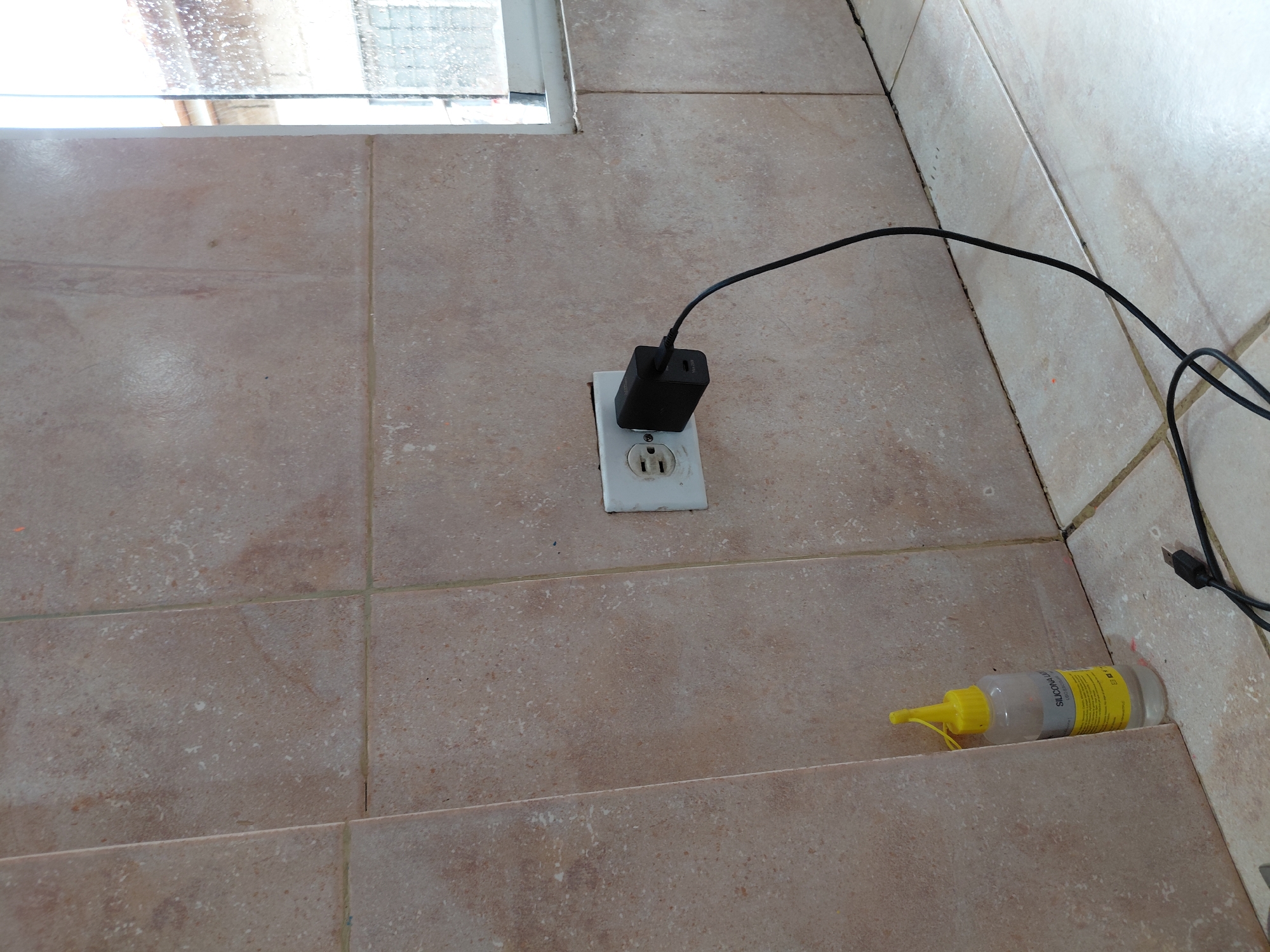 The image captures a ground-level view of a tiled area, possibly indoors or a covered outdoor space. The tiles are light brown or beige, square-shaped, with a textured, somewhat rustic appearance and visible grout lines.
In the mid-left portion of the frame, a white electrical outlet is mounted into the tiled surface. A black wall charger is plugged into the upper socket of the outlet. A black electrical cable extends from the charger, running across the tiled floor towards the right side of the image and out of view, suggesting it connects to a device not visible in the frame. The outlet and adjacent tiles exhibit some dirt or wear.
In the top left, a portion of a window is visible, letting in natural light. The view through the window is somewhat blurred, showing light-colored, possibly textured surfaces or reflections.
Towards the lower right, on what appears to be a lower step or floor level, a clear plastic bottle with a bright yellow cap and nozzle is lying horizontally. The bottle contains a clear liquid. Text visible on the bottle's label reads "SILICONA LIQUIDA", indicating it is liquid silicone. Other smaller text on the label is present but not legible.
The scene depicts a functional, utilitarian space, possibly a utility room, kitchen, or a patio area in Santa Ana, Venezuela, during daylight hours. No people are visible, and there is no discernible activity beyond the charger being plugged in.