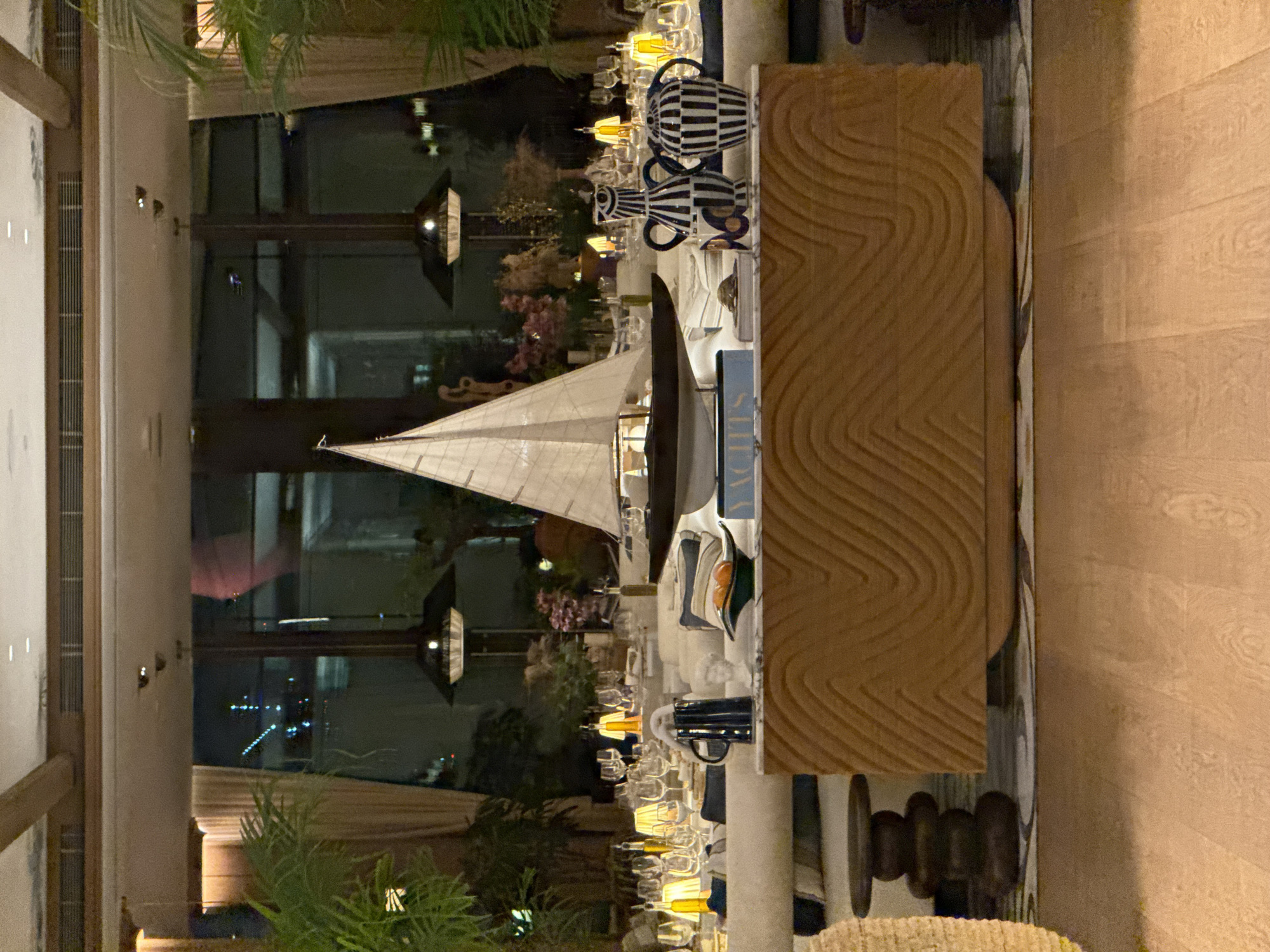 The image shows an interior setting, likely a restaurant or event space, in Dubai, United Arab Emirates, based on the presence of waterfront views and distinctive architectural styles often found in that city. The composition is somewhat vertically oriented, with the main focus on a dining setup featuring a centerpiece of a model sailboat. Other decor includes striped vases, floral arrangements, and glassware, suggesting a formal dining experience. The background reveals a cityscape through a large window, hinting at a night scene. The light is artificial and warm, contributing to an elegant atmosphere. The room seems to be well-appointed and designed for upscale events.